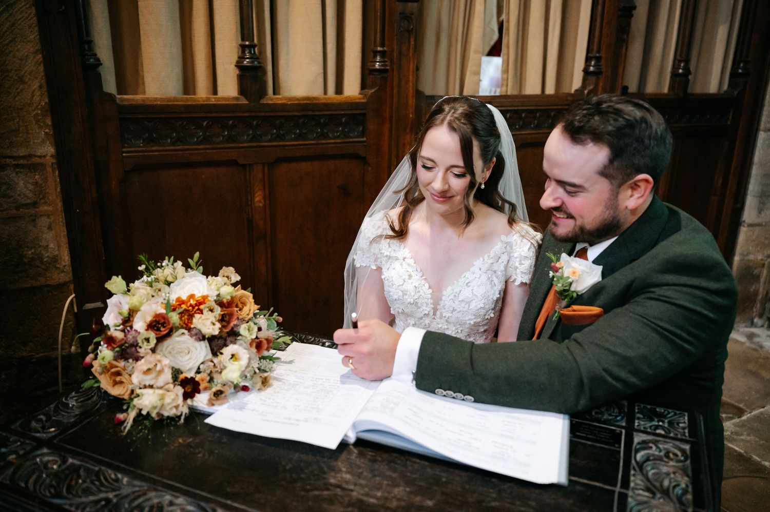 Bride in a lace gown and veil smiles as the groom signs the wedding register beside a bouquet of peach and cream flowers.