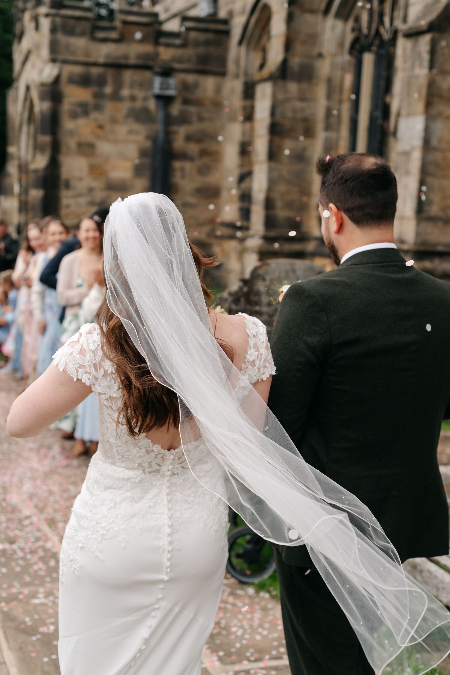 Bride in a long flowing veil and groom in a dark suit walk past cheering guests as confetti falls outside a stone church.