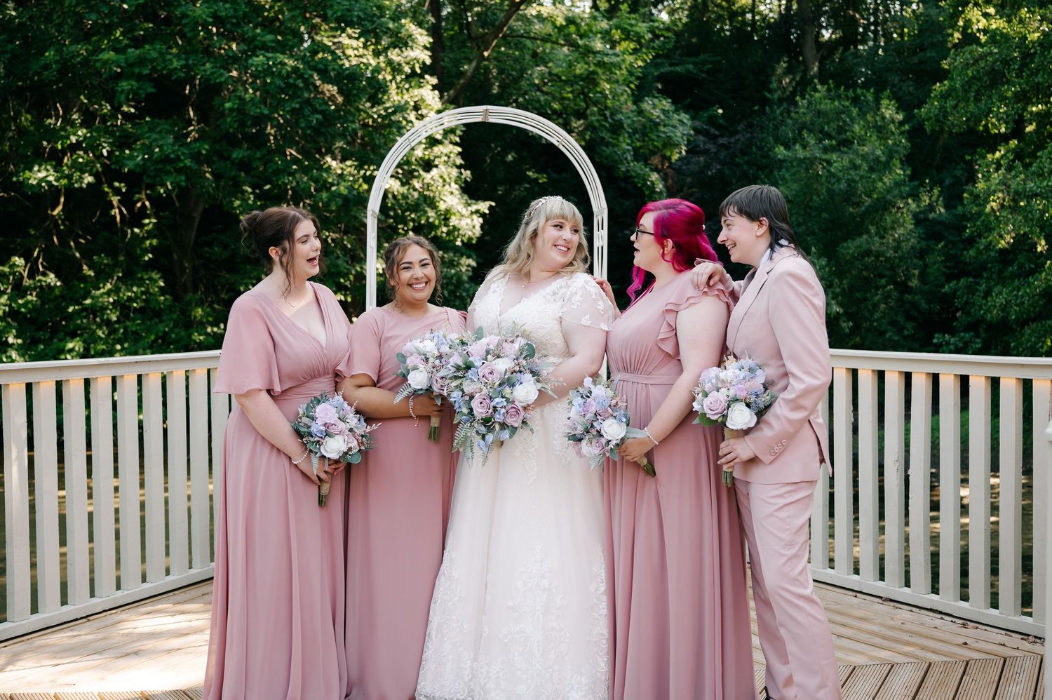 Bride in white lace gown poses with bridesmaids in dusty rose dresses and suit on wooden deck.