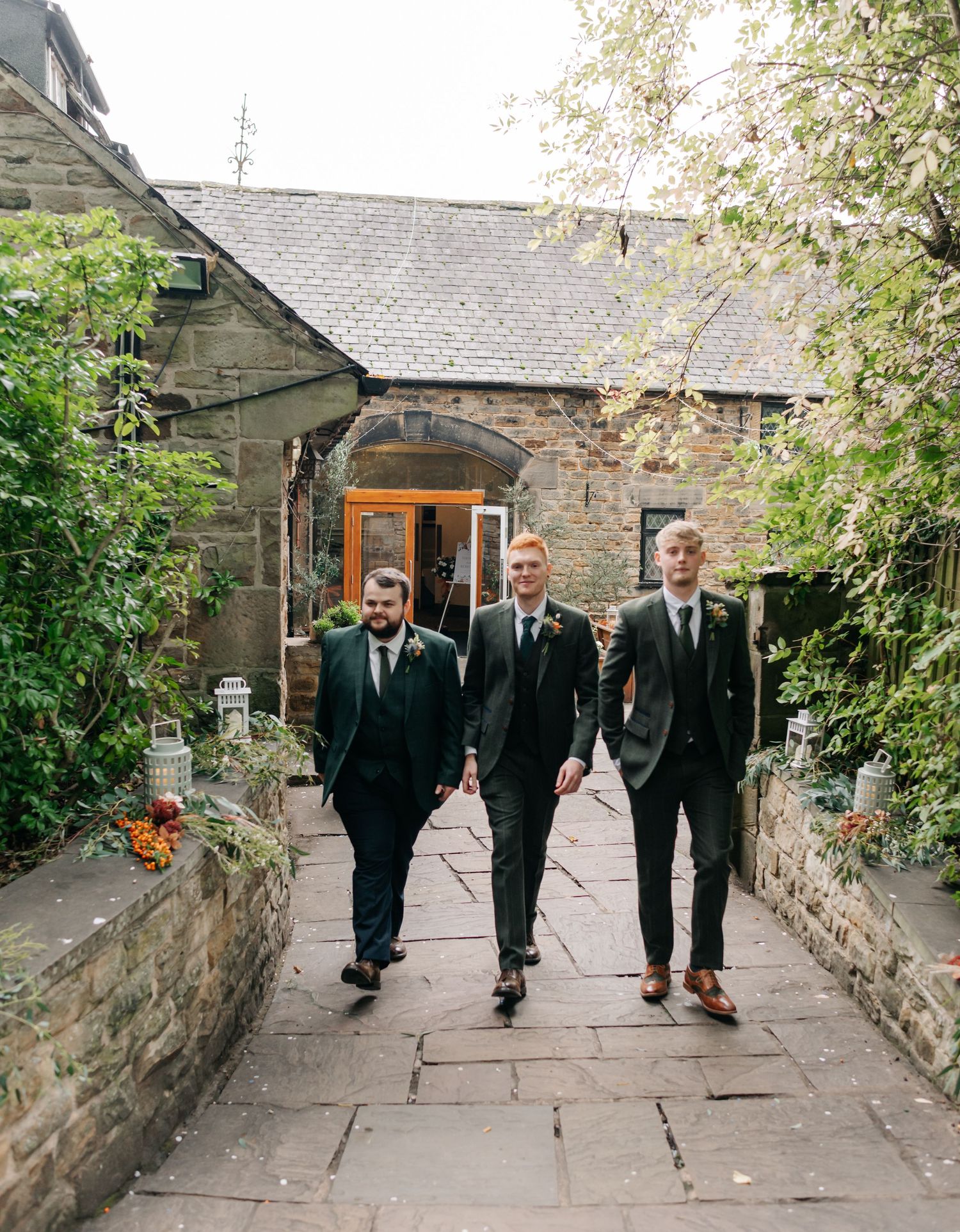 Three groomsmen walking along a stone path outside a rustic building.