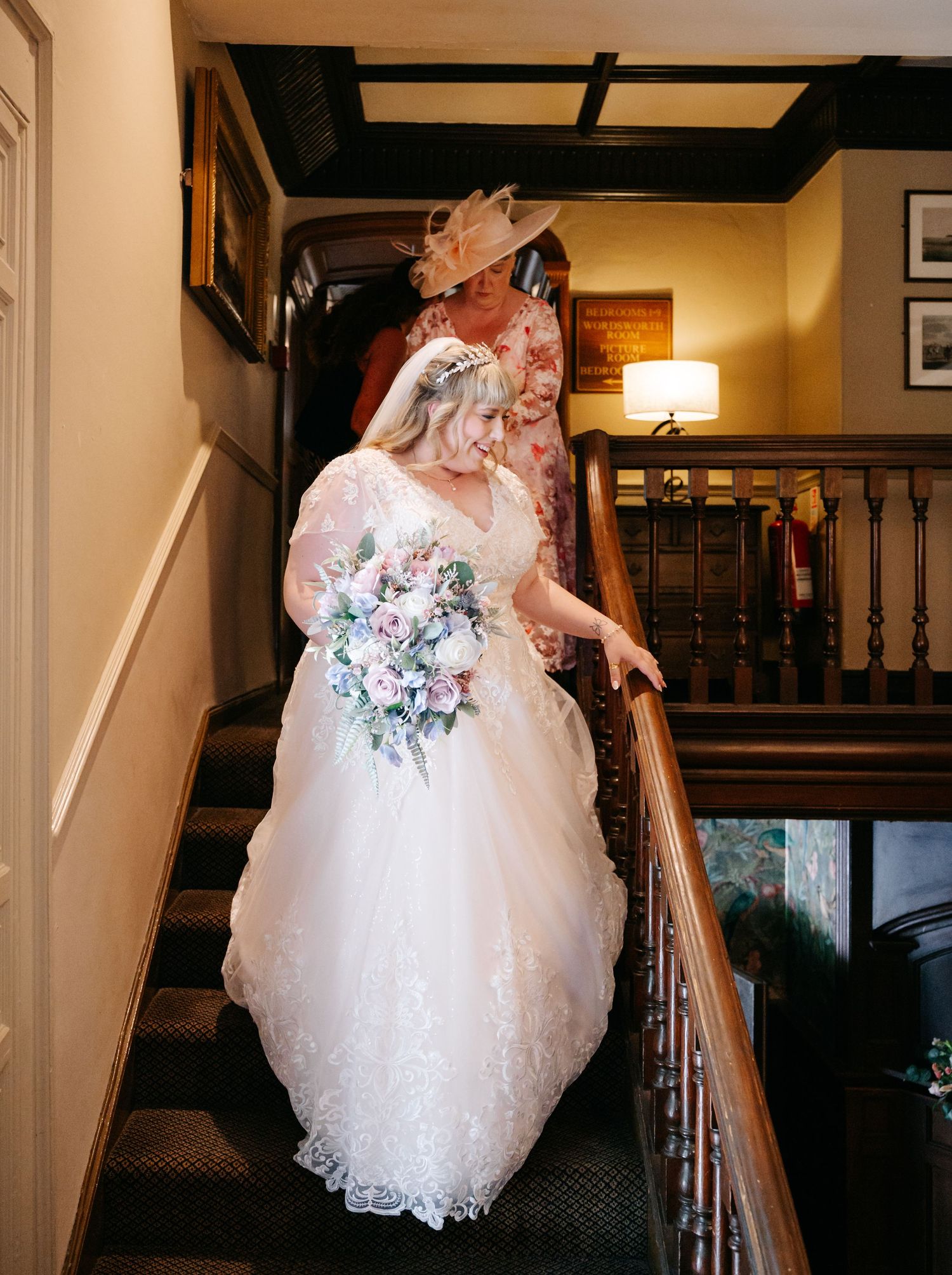 Beautiful bride in lace ballgown with floral bouquet descending staircase accompanied by guest in pink hat.
