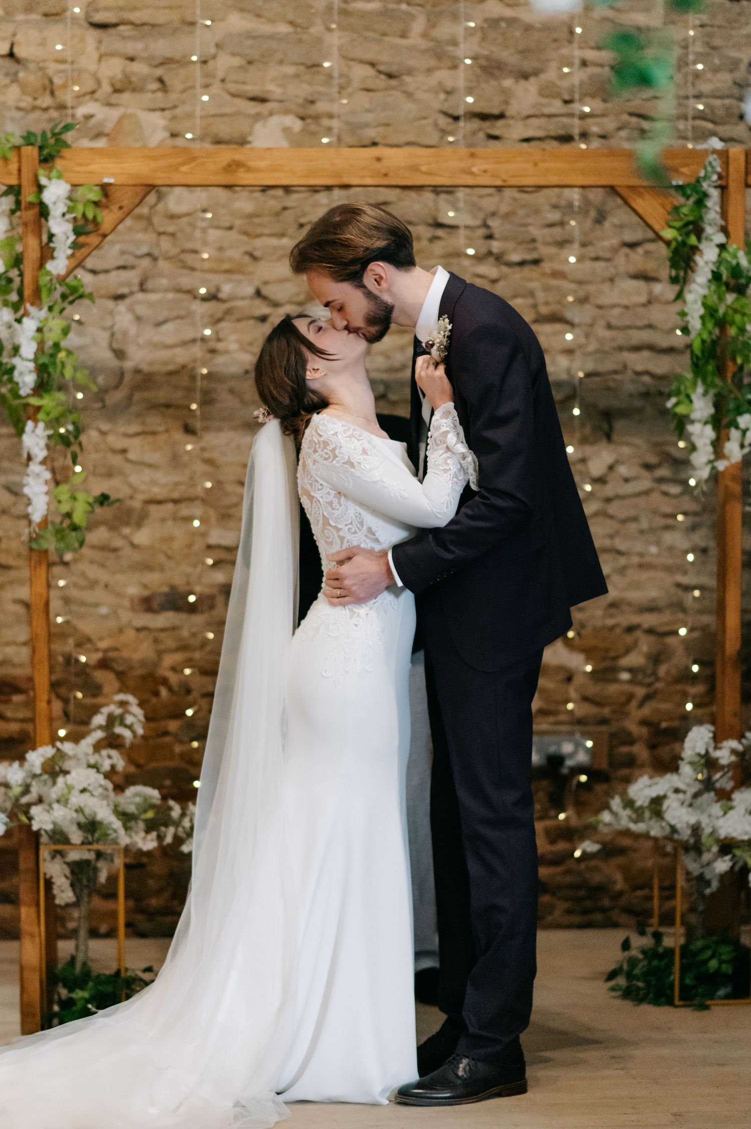 A romantic wedding ceremony moment under a wooden arch adorned with greenery and string lights.