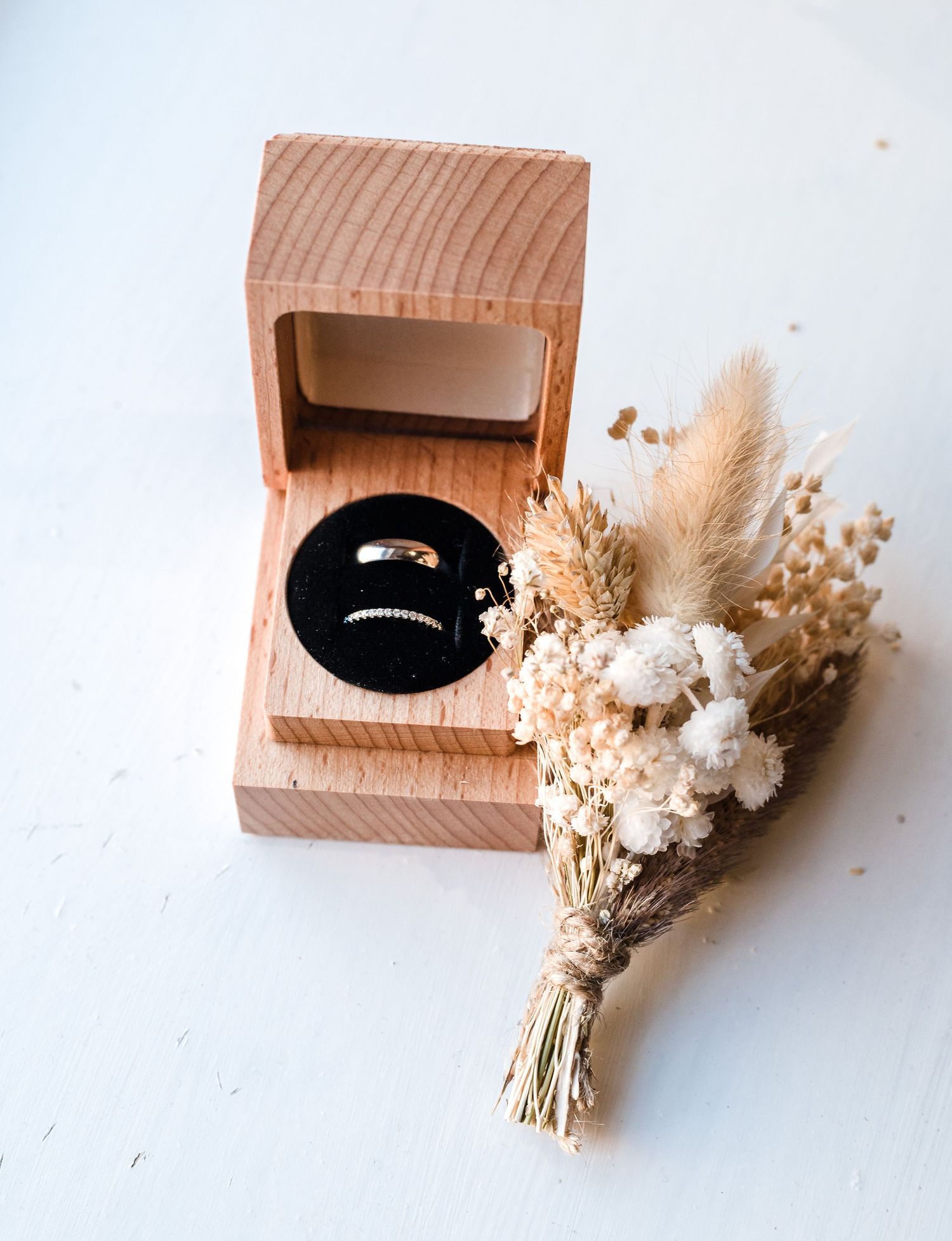 Wooden ring box with wedding bands displayed alongside dried flower bouquet.