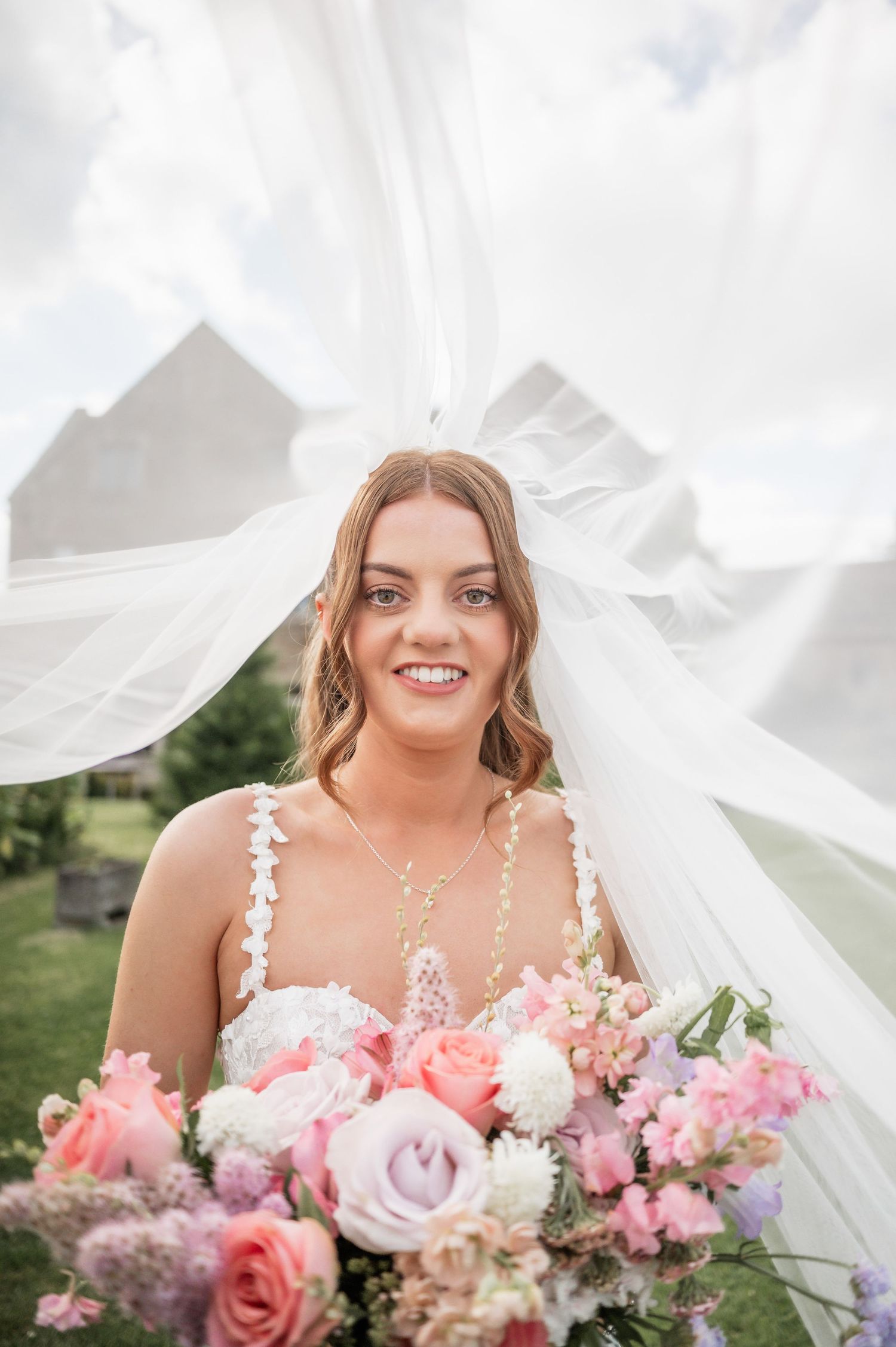 Smiling bride with flowing veil holds romantic pastel bouquet outdoors.