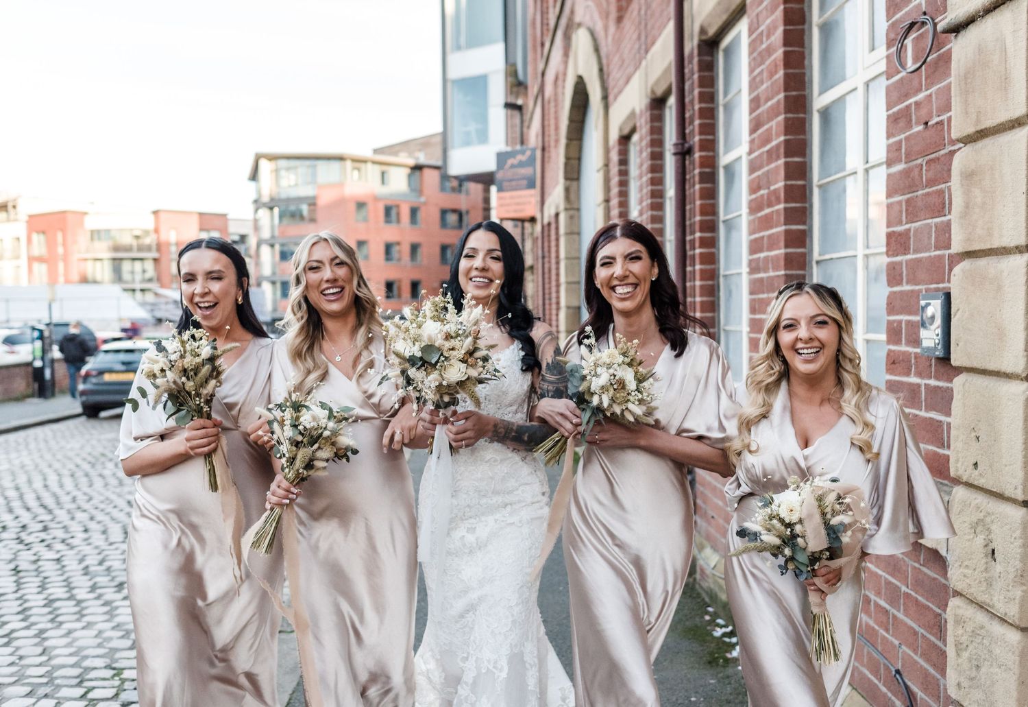 Bridesmaids in blush pink dresses walk down a cobblestone street carrying neutral floral bouquets.