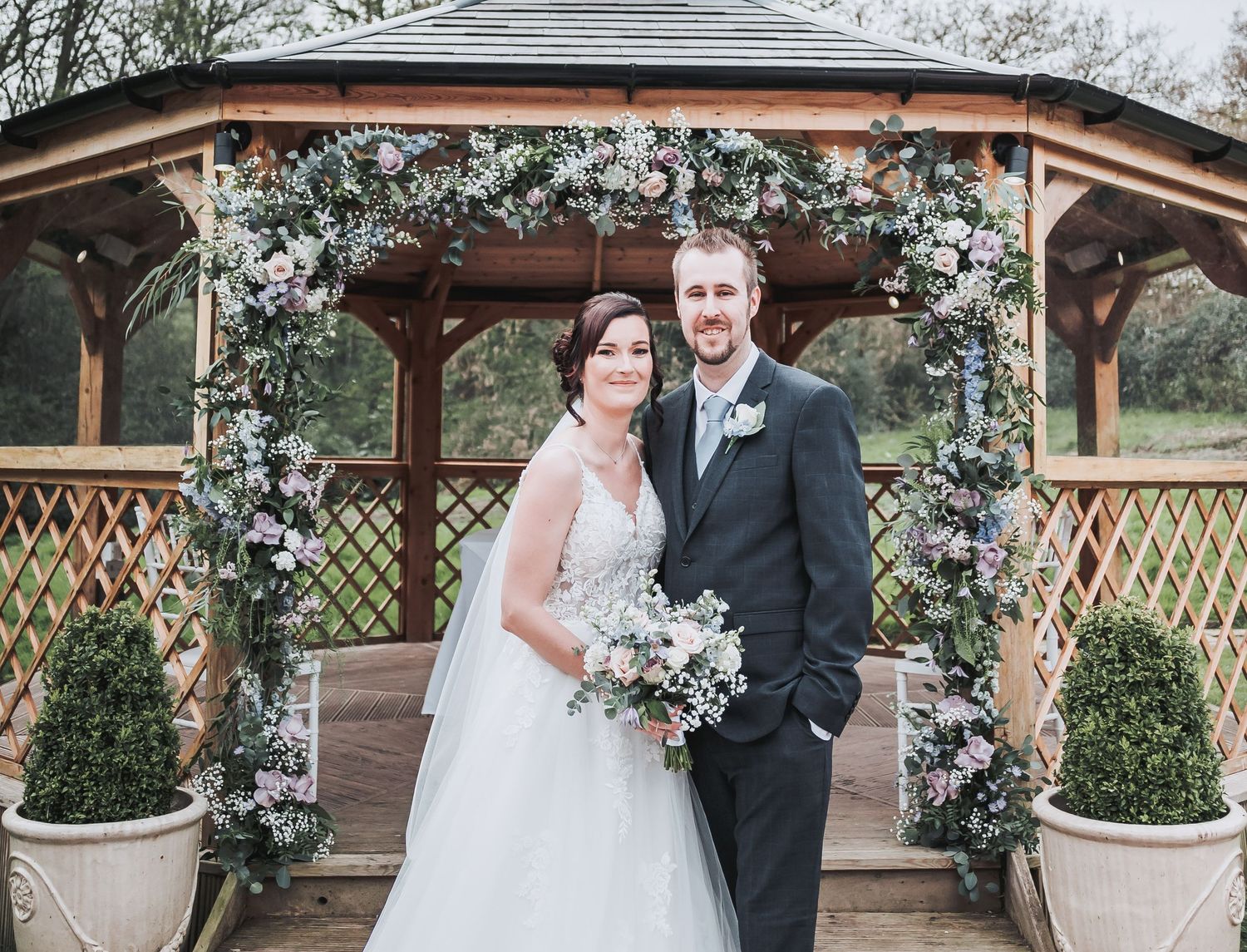 A wedding couple poses under a decorated wooden gazebo with floral arrangements.