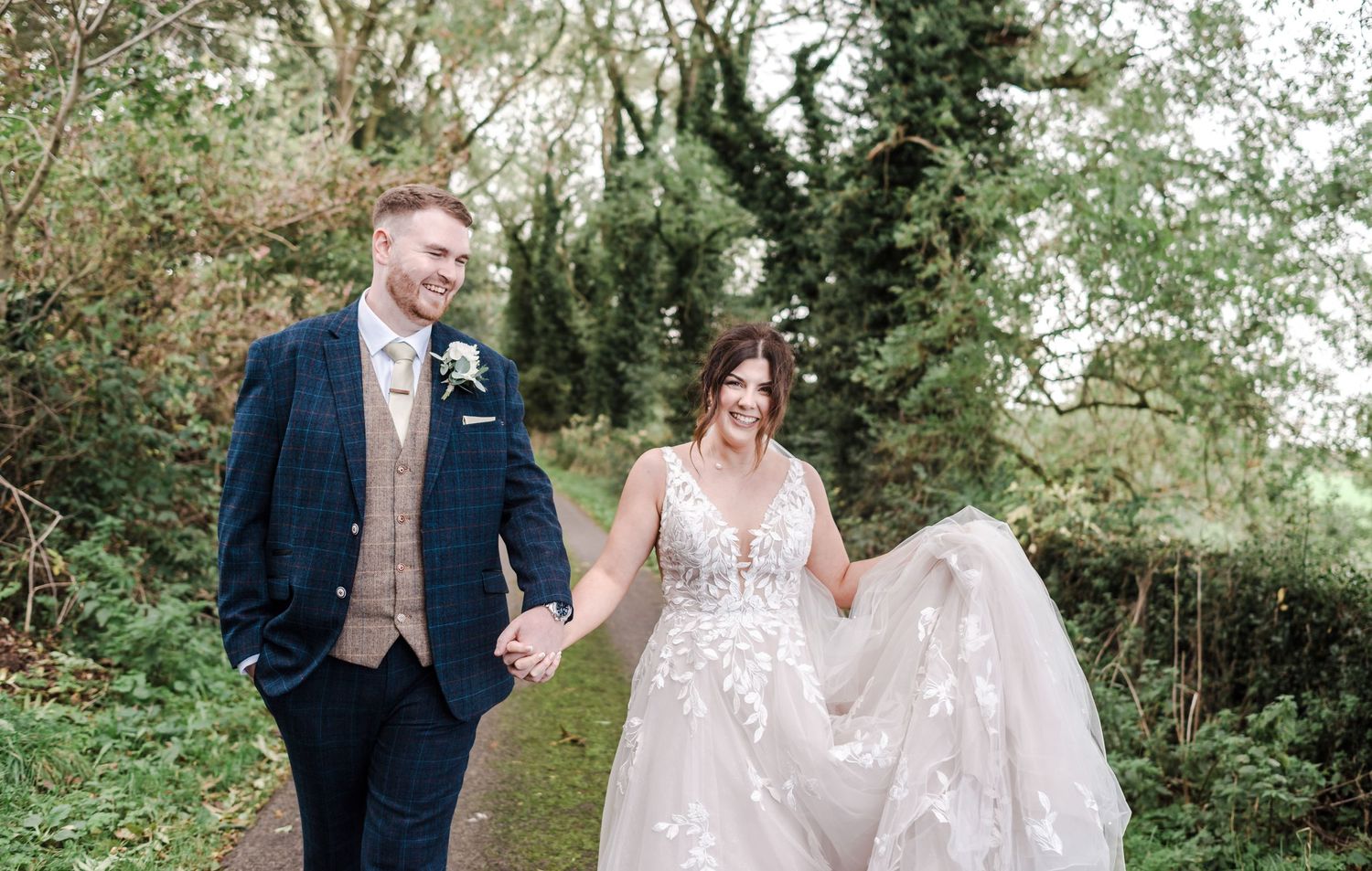 Couple walking on a tree-lined path with flowing white dress and navy suit for outdoor wedding photography.