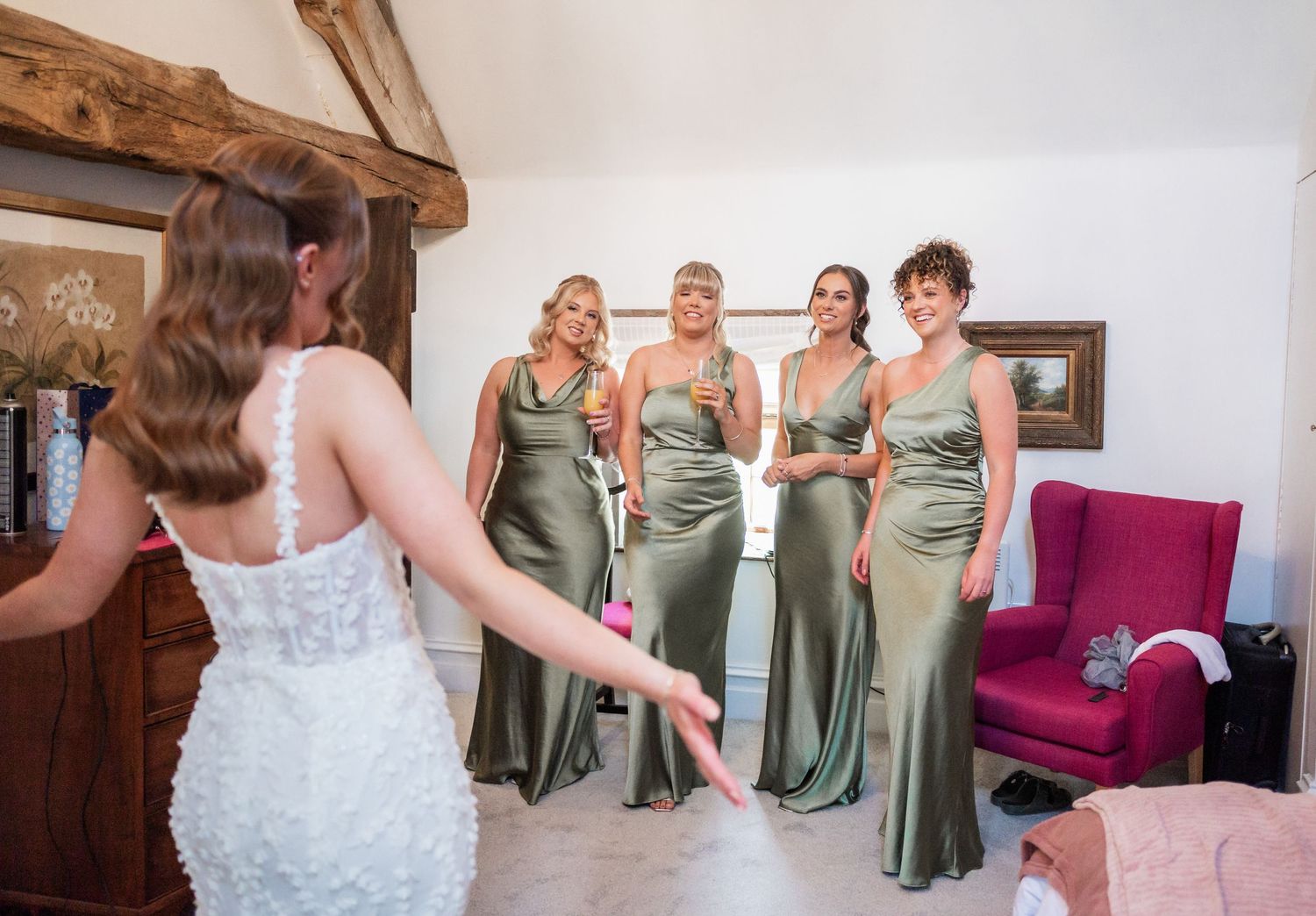 Bridesmaids in sage green dresses react to bride's dress reveal in rustic room.