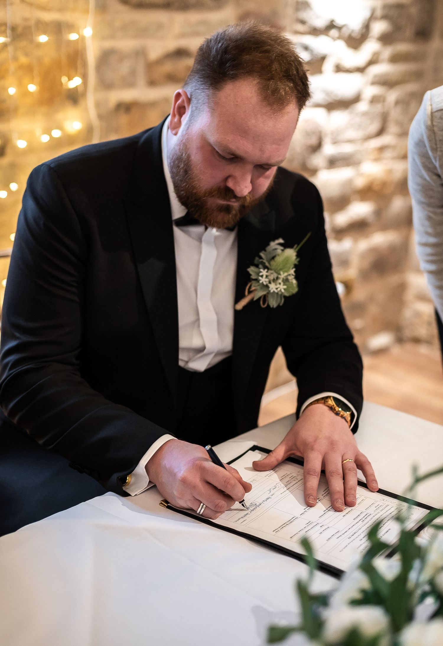 Person in tuxedo signing wedding documents at decorated table with white florals.