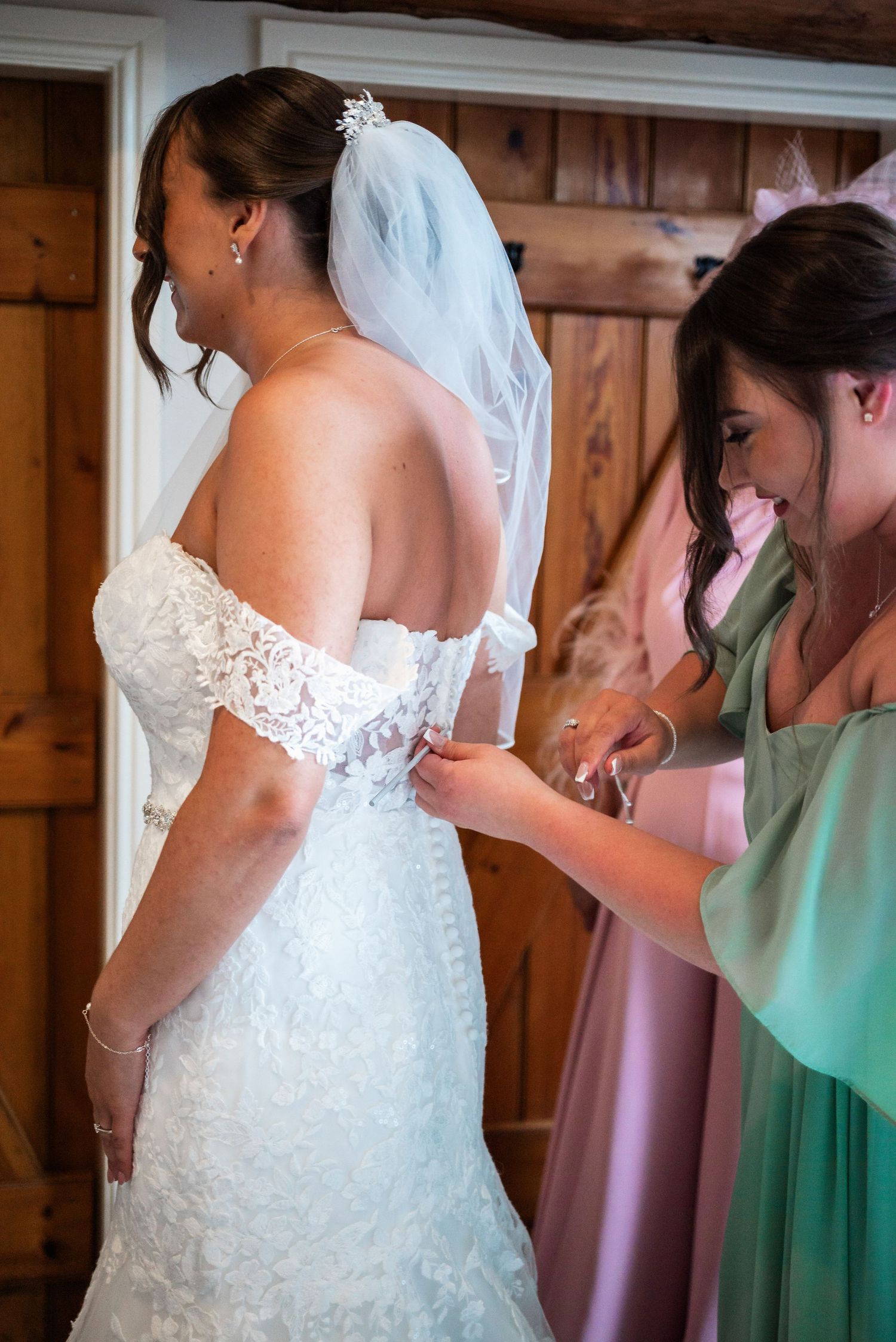 A bridesmaid in mint green helps adjust the back of a white lace wedding dress during preparation.