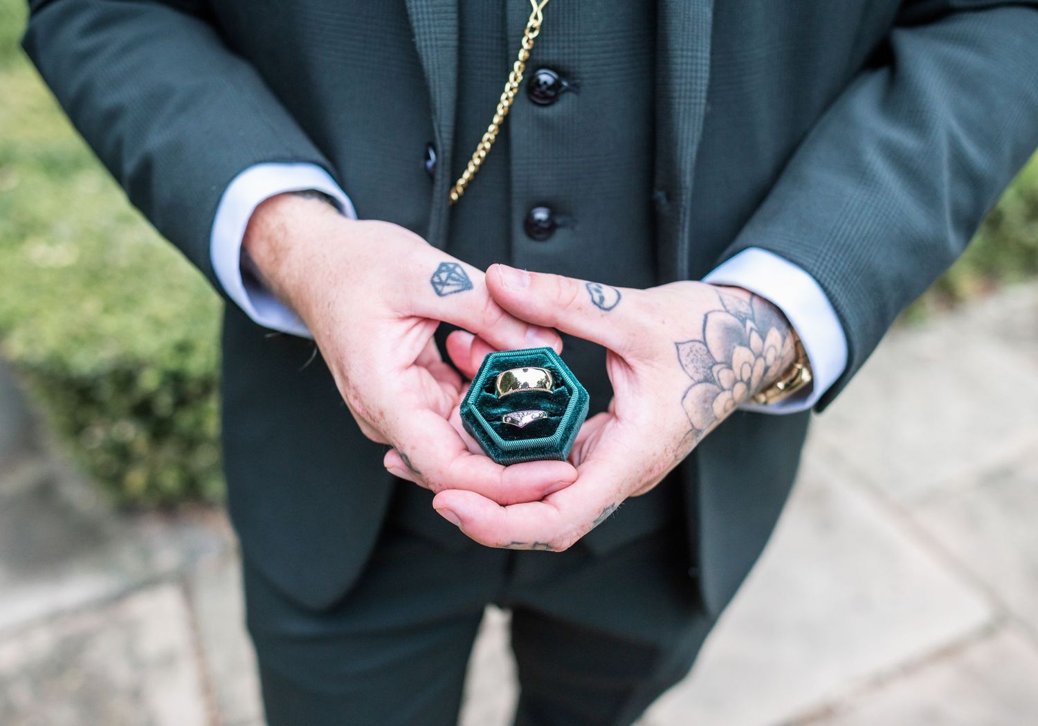 Tattooed hands holding emerald green hexagonal ring box showing wedding bands.