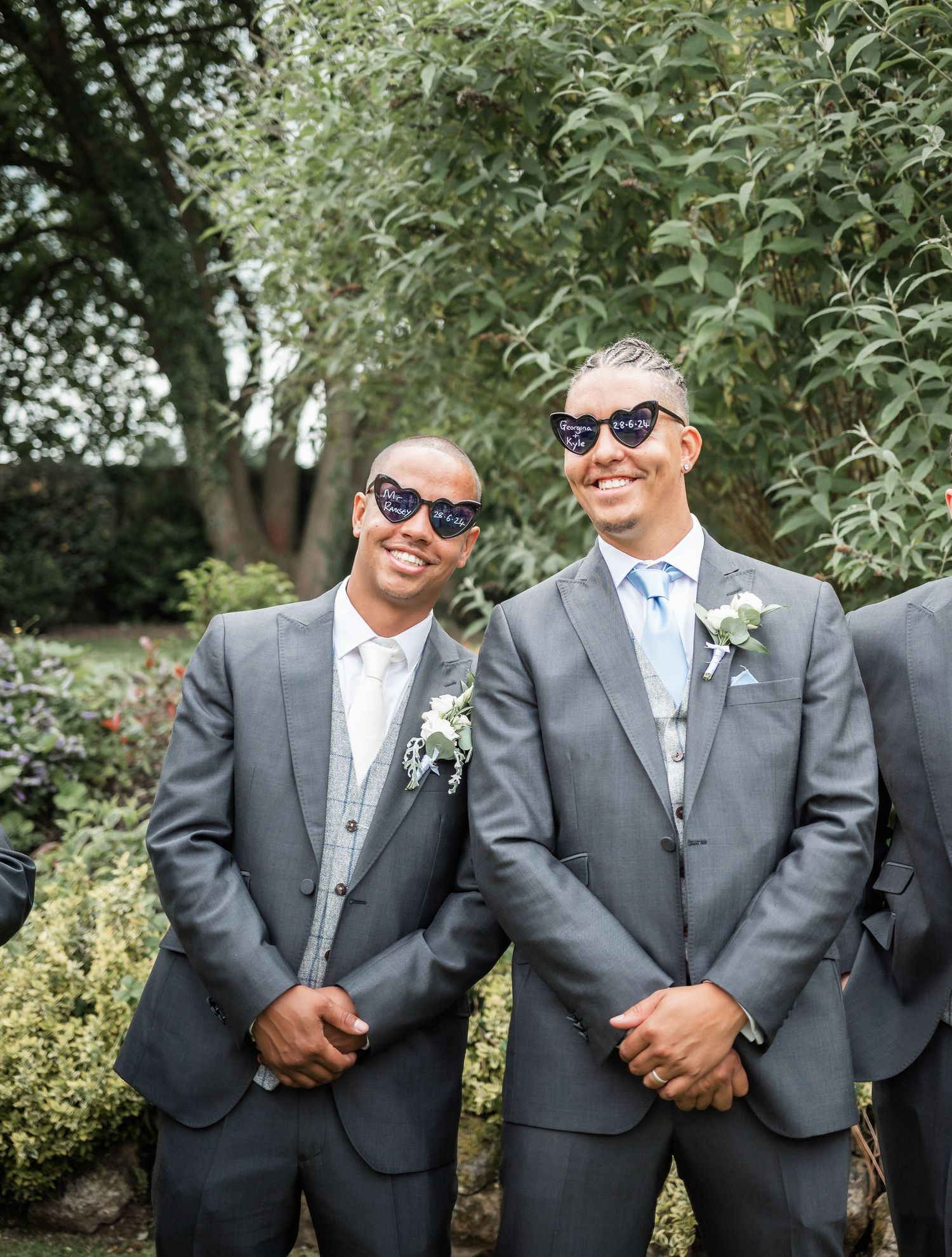 Two groomsmen in matching grey suits and sunglasses smile outdoors in a garden setting.