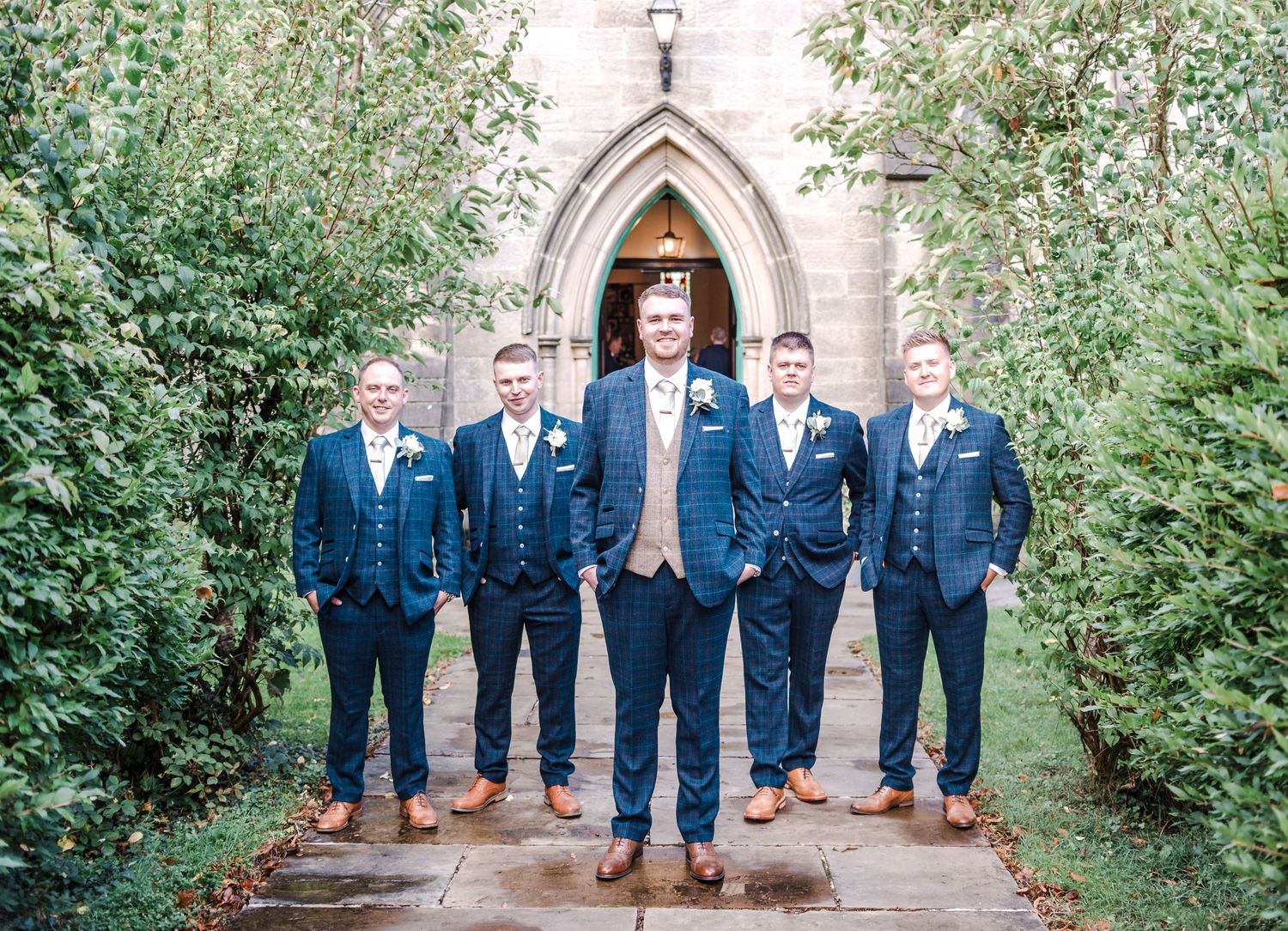 Groomsmen in navy suits pose at church entrance framed by greenery.