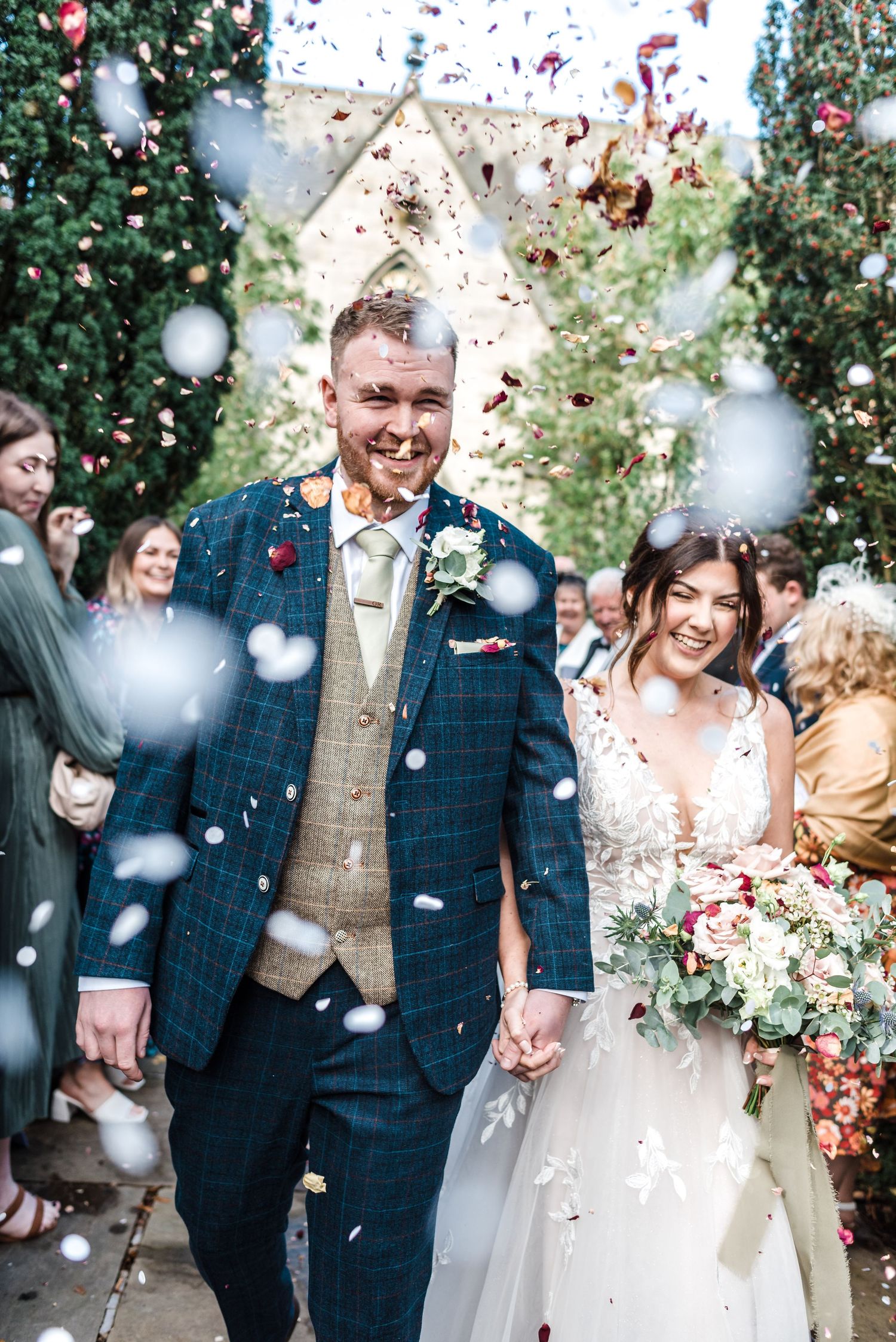 Wedding party smiling outdoors surrounded by trees and confetti.