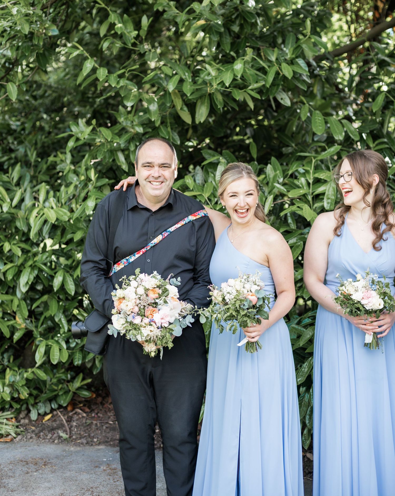 Wedding party members in black and light blue attire holding floral bouquets against green foliage.