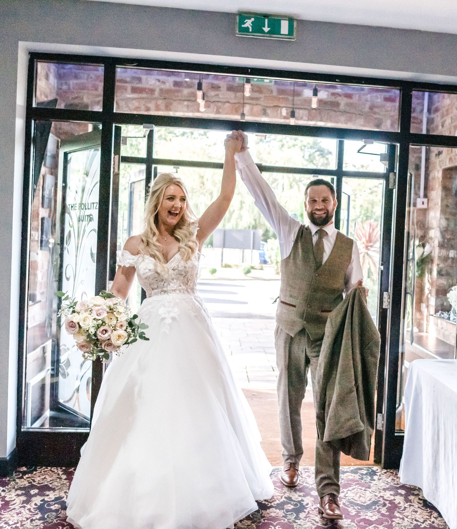 Newlyweds make their grand entrance through glass doors as the bride holds a bouquet and groom wears a tan vest.