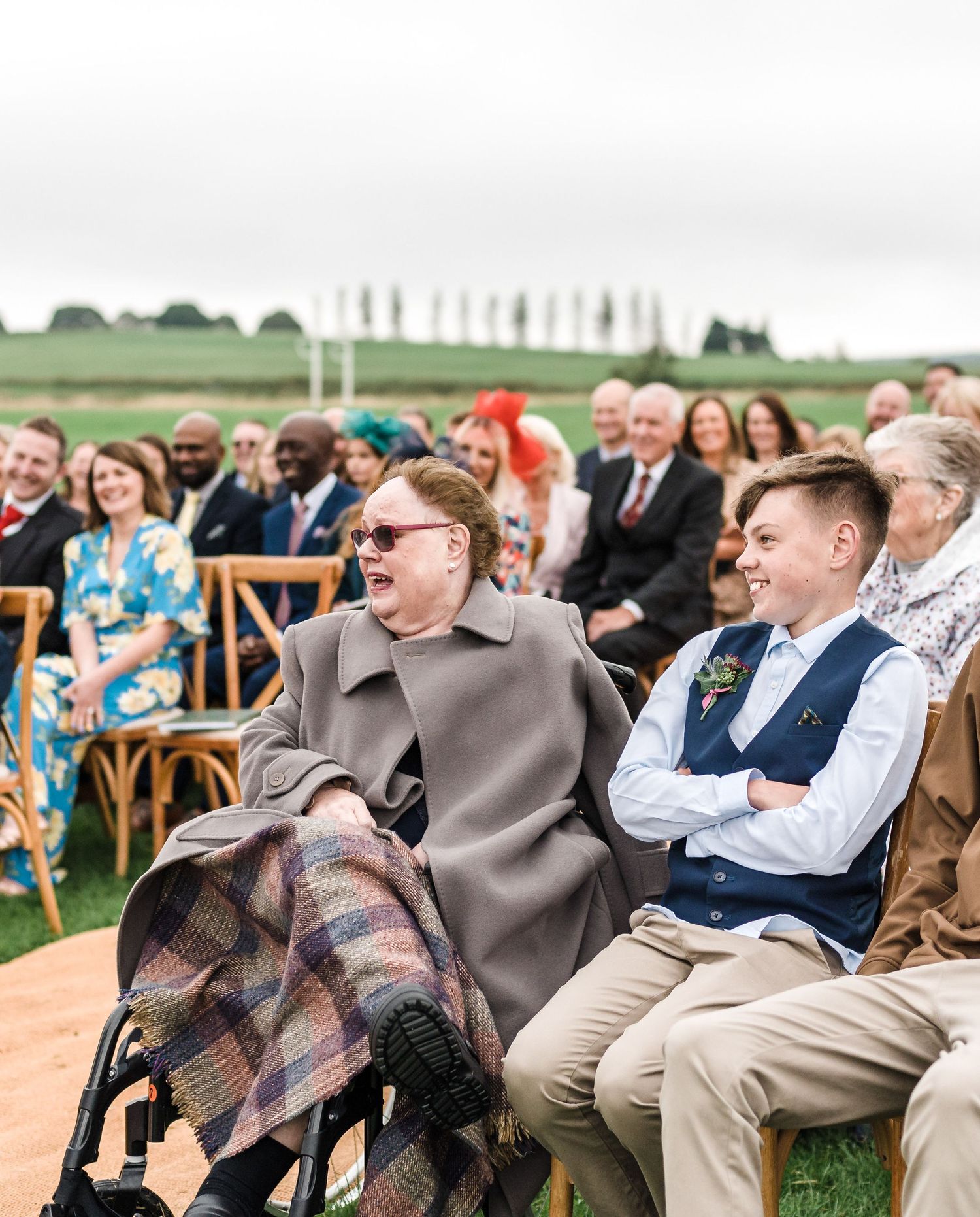 Wedding guests seated in wooden chairs watching ceremony in outdoor countryside setting.