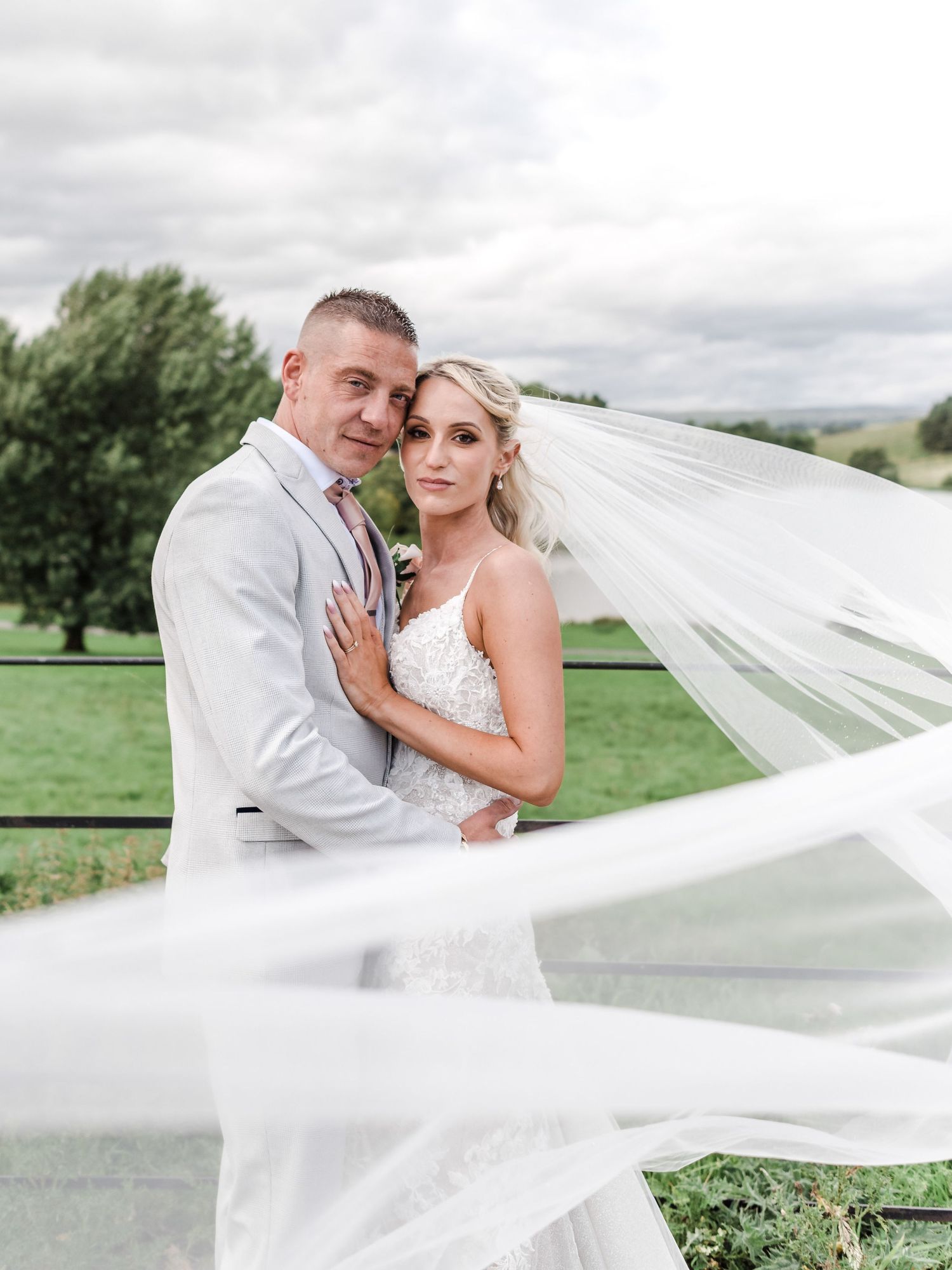 Romantic wedding portrait with flowing veil against scenic countryside background.