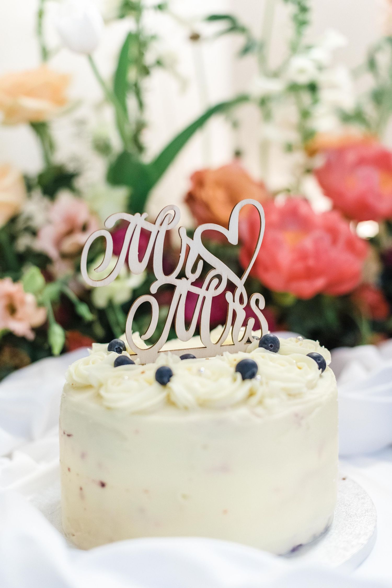 A white wedding cake topped with a silver 'Mr & Mrs' topper and fresh blueberries against floral backdrop.