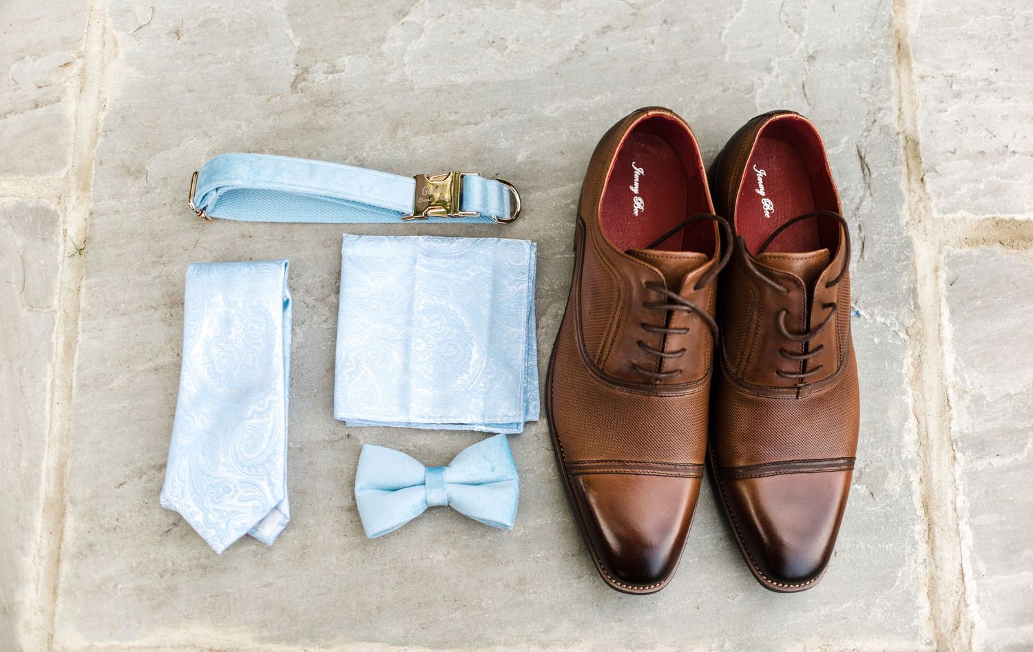 Wedding accessories including a light blue tie, bowtie, and brown leather dress shoes arranged on stone.