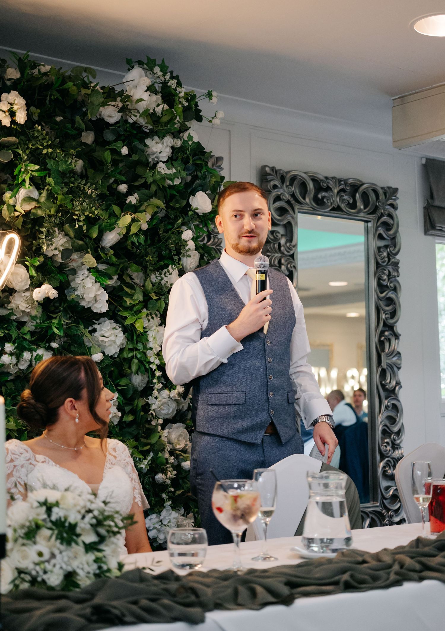 Wedding reception scene with floral decorations and speeches at an elegant indoor venue.
