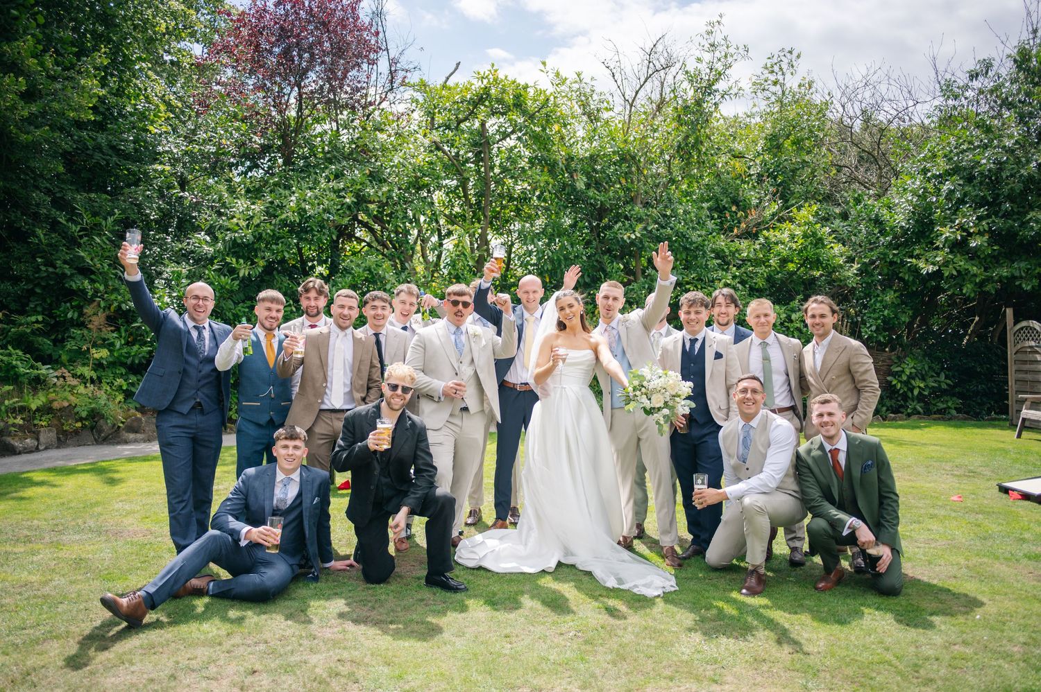 Large wedding party poses for group photos on lawn with trees in background.
