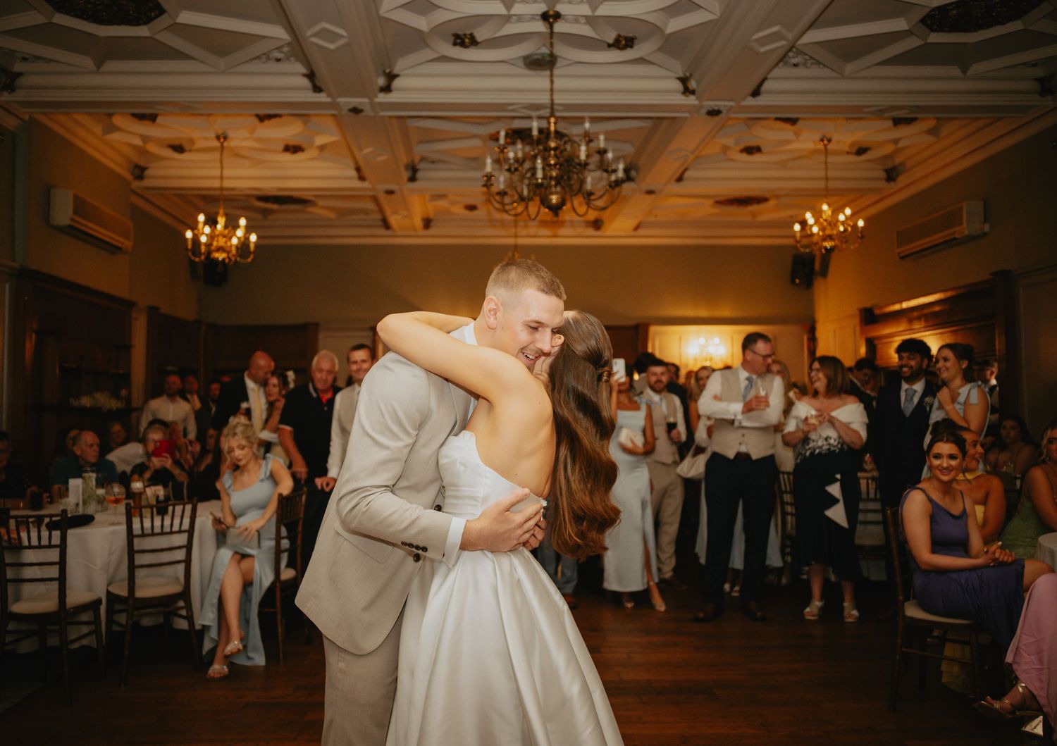 Wedding couple shares their first dance in a dimly lit ballroom with chandelier.