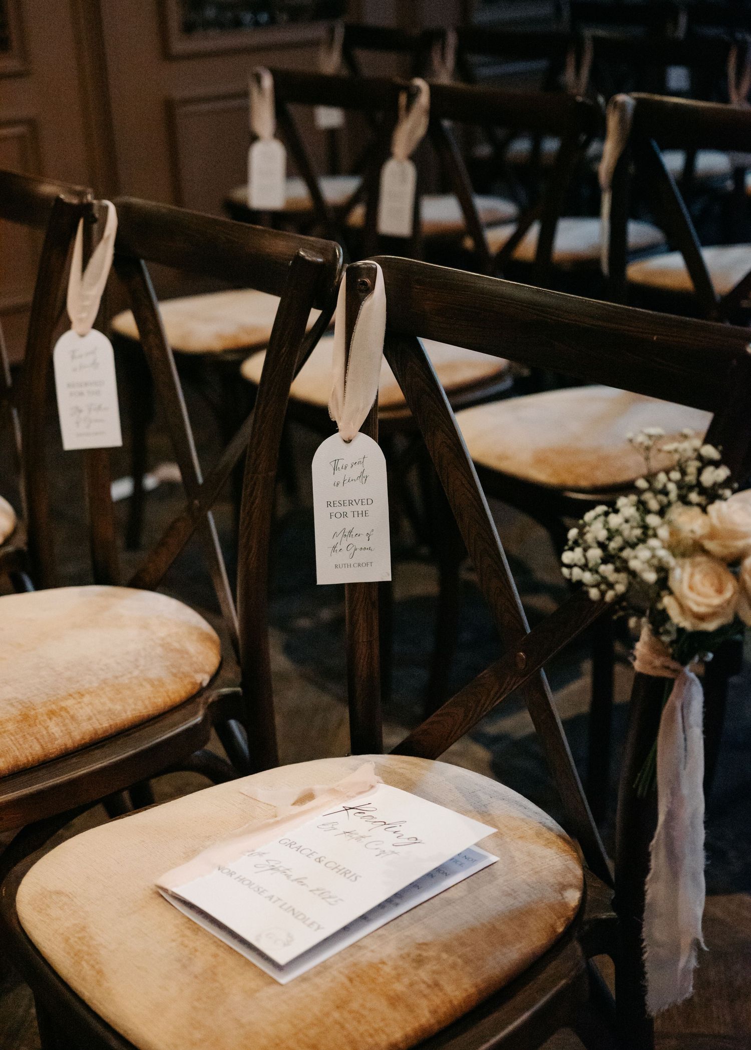 Reserved seating cards with white ribbons hang on dark wooden chairs at an elegant wedding ceremony setup.