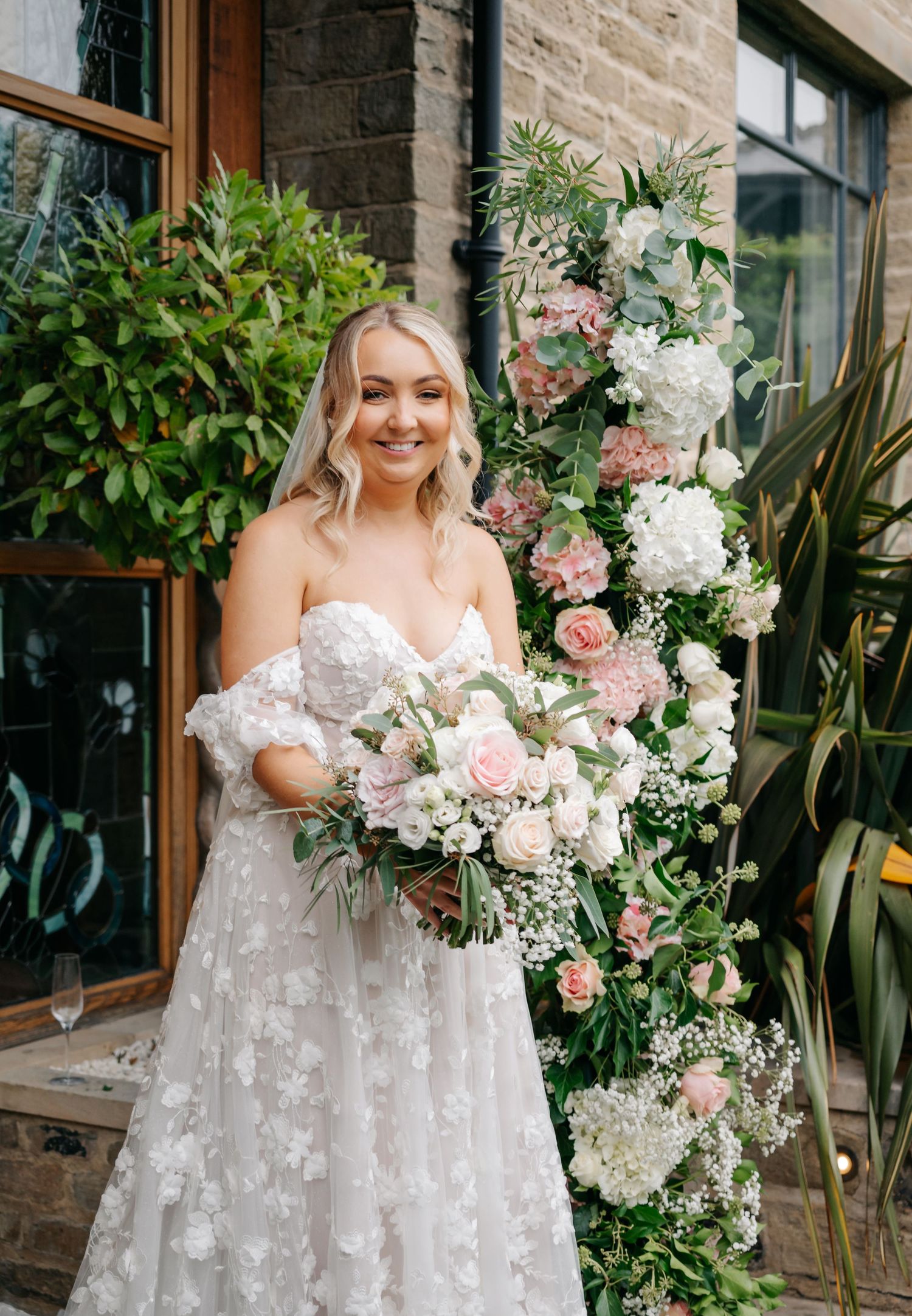 Bride in detailed white gown holds pink and white floral bouquet against stone wall backdrop.
