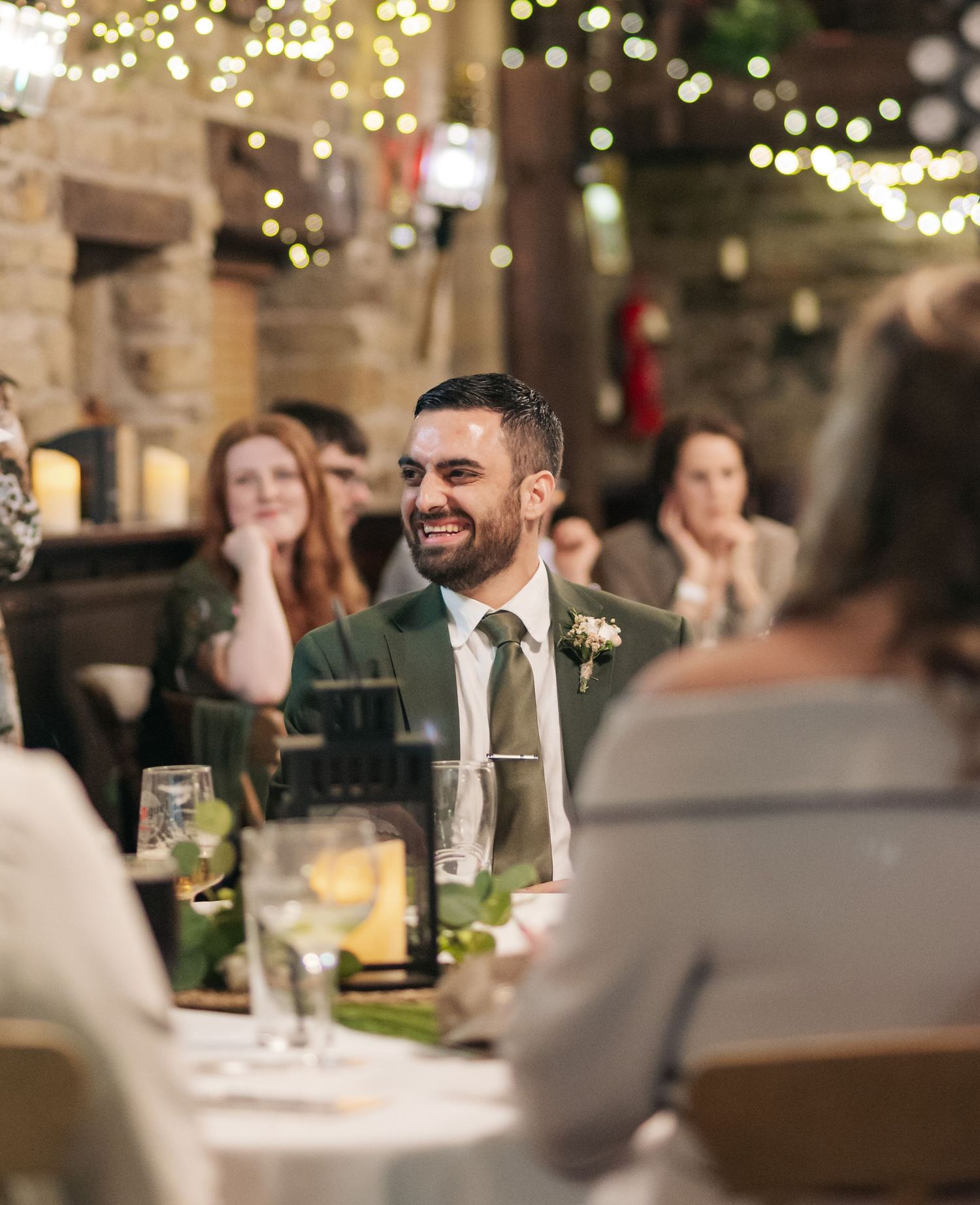 Wedding guests seated at dinner tables with twinkling lights and stone walls in the background.