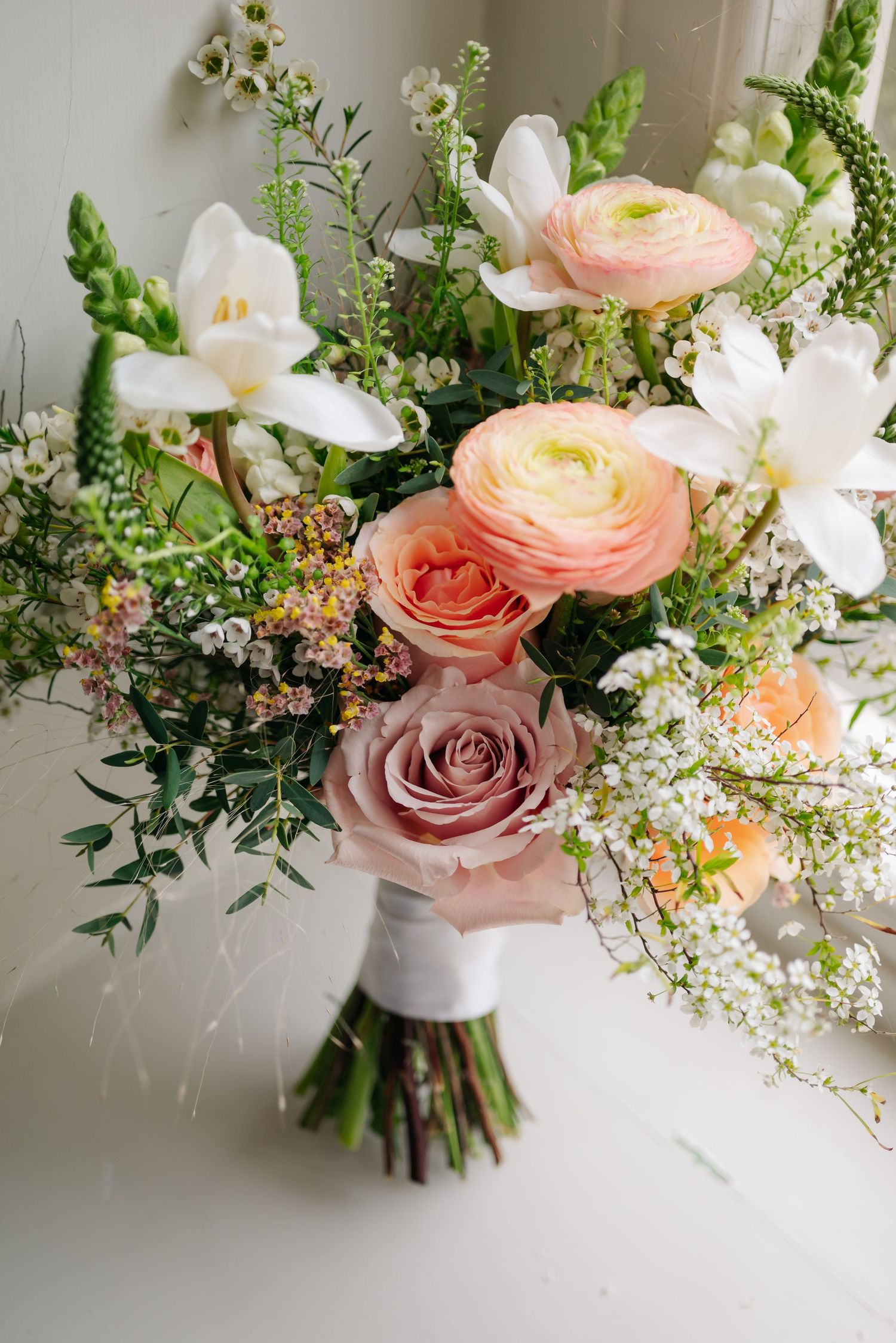 Elegant wedding bouquet featuring blush roses, ranunculus, and delicate greenery against a light background.