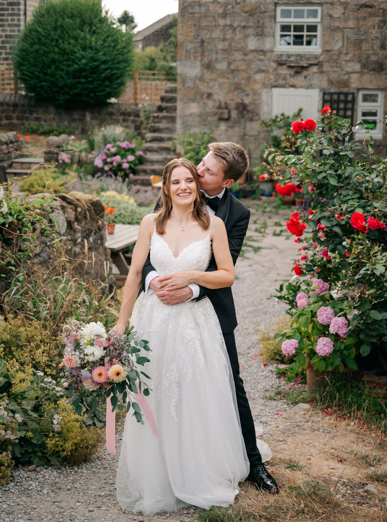 Bride in a lace wedding dress holding a bouquet smiles as the groom embraces her from behind in a blooming stone-walled garden.