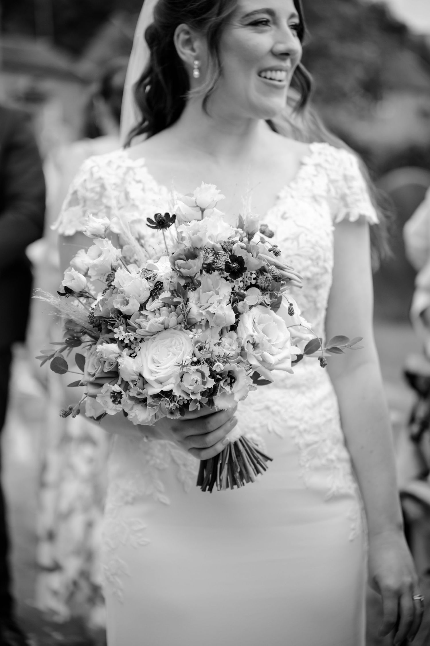 Smiling bride in a lace cap-sleeve wedding dress holds a romantic bouquet of roses and wildflowers in a black and white photo.