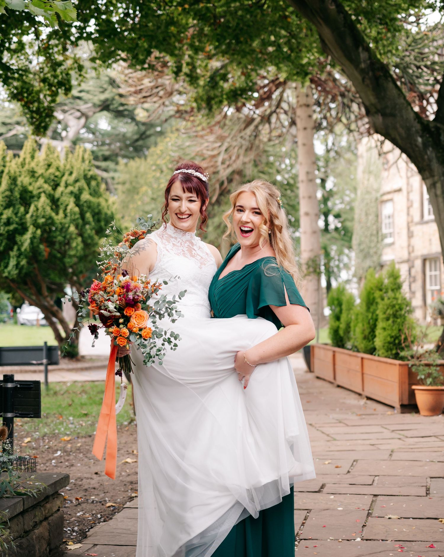Two women share a joyful moment on stone pathway during wedding celebrations.