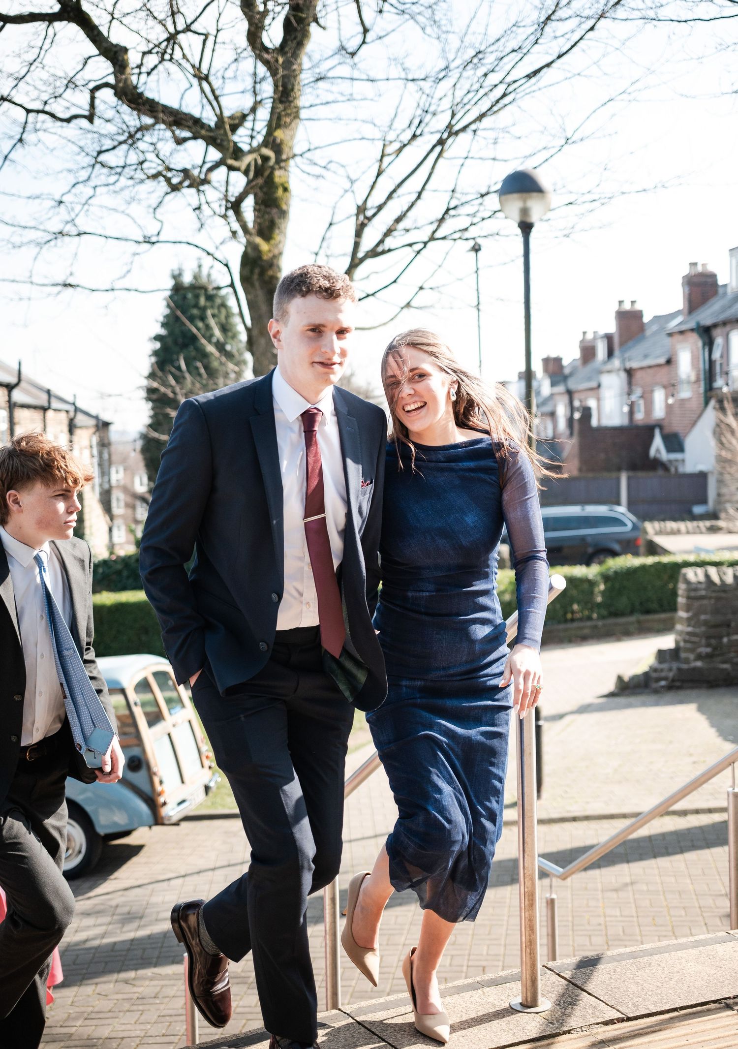 A couple walking together outdoors, with the woman wearing a navy dress and the man in a dark suit and tie.