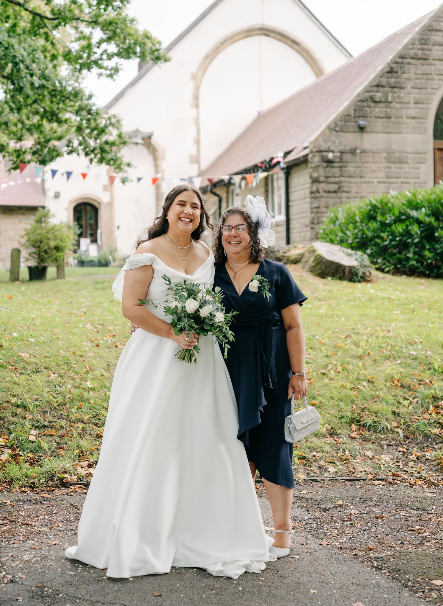 Wedding guests pose together on a grassy lawn outside a church with white stone walls and arched doorway.