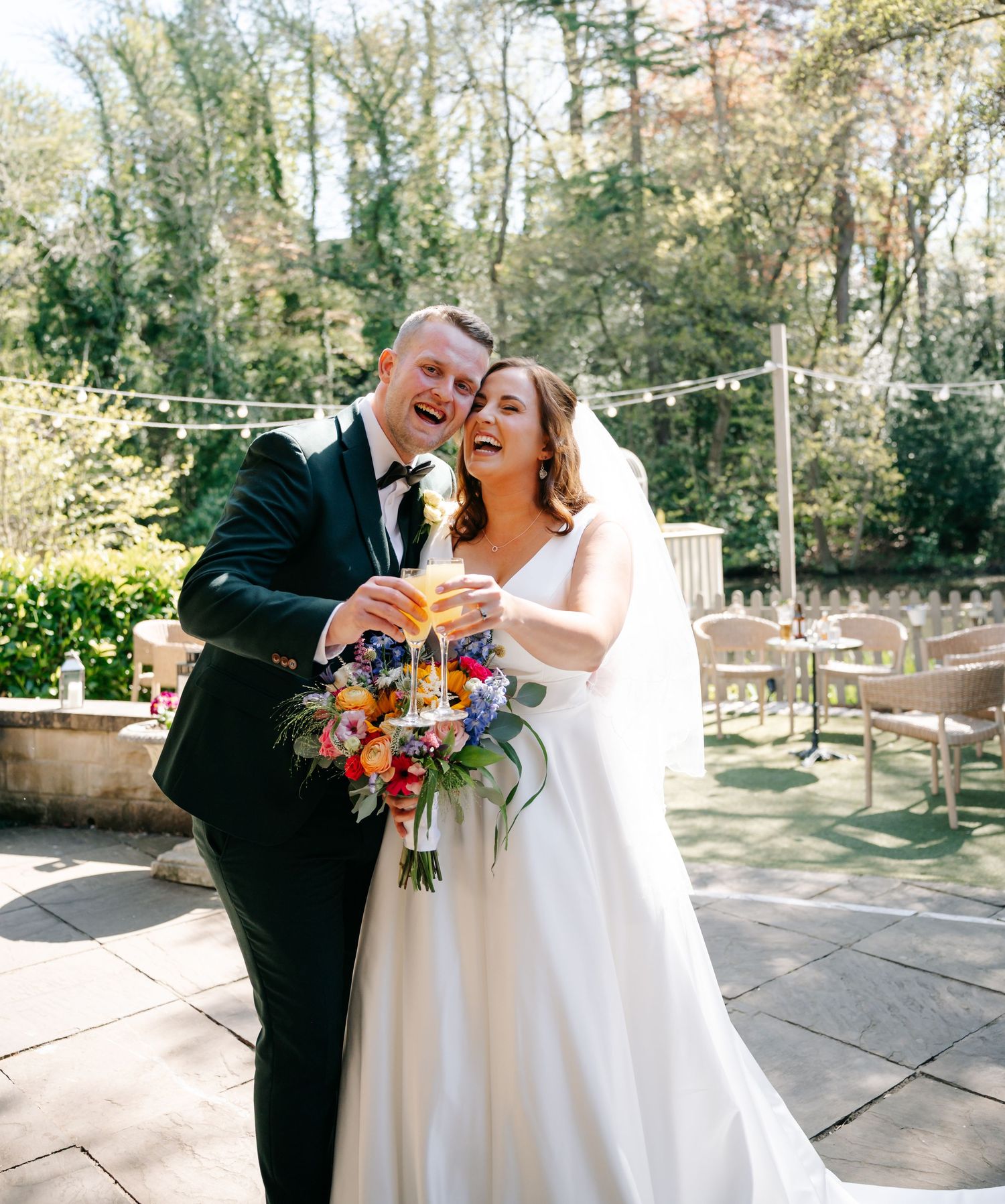 Happy newlyweds toasting with champagne, bride holding colorful bouquet in white gown, groom in black tuxedo, outdoor garden venue.