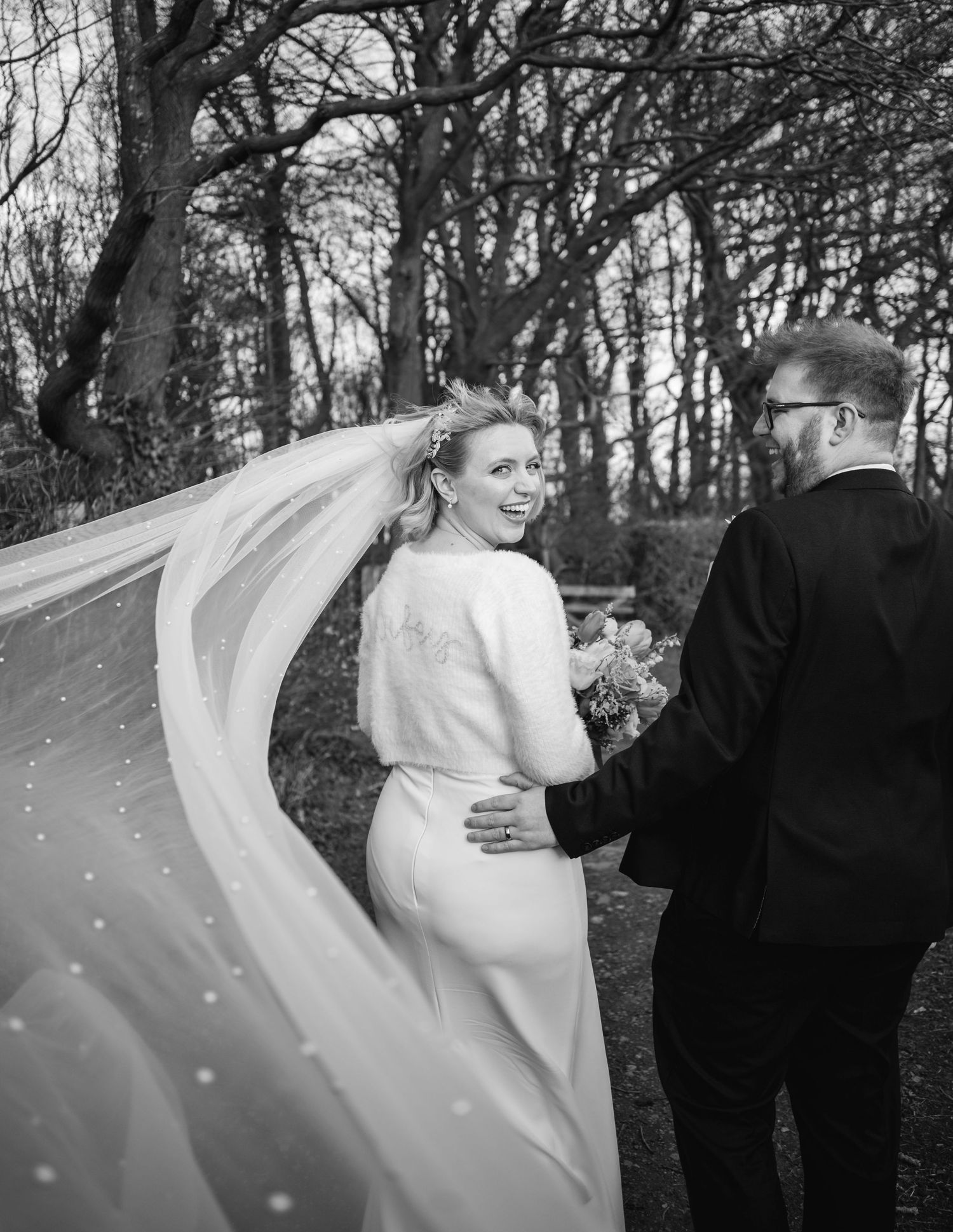 Black and white photo of a smiling bride with flowing veil walking with groom outdoors among bare trees.