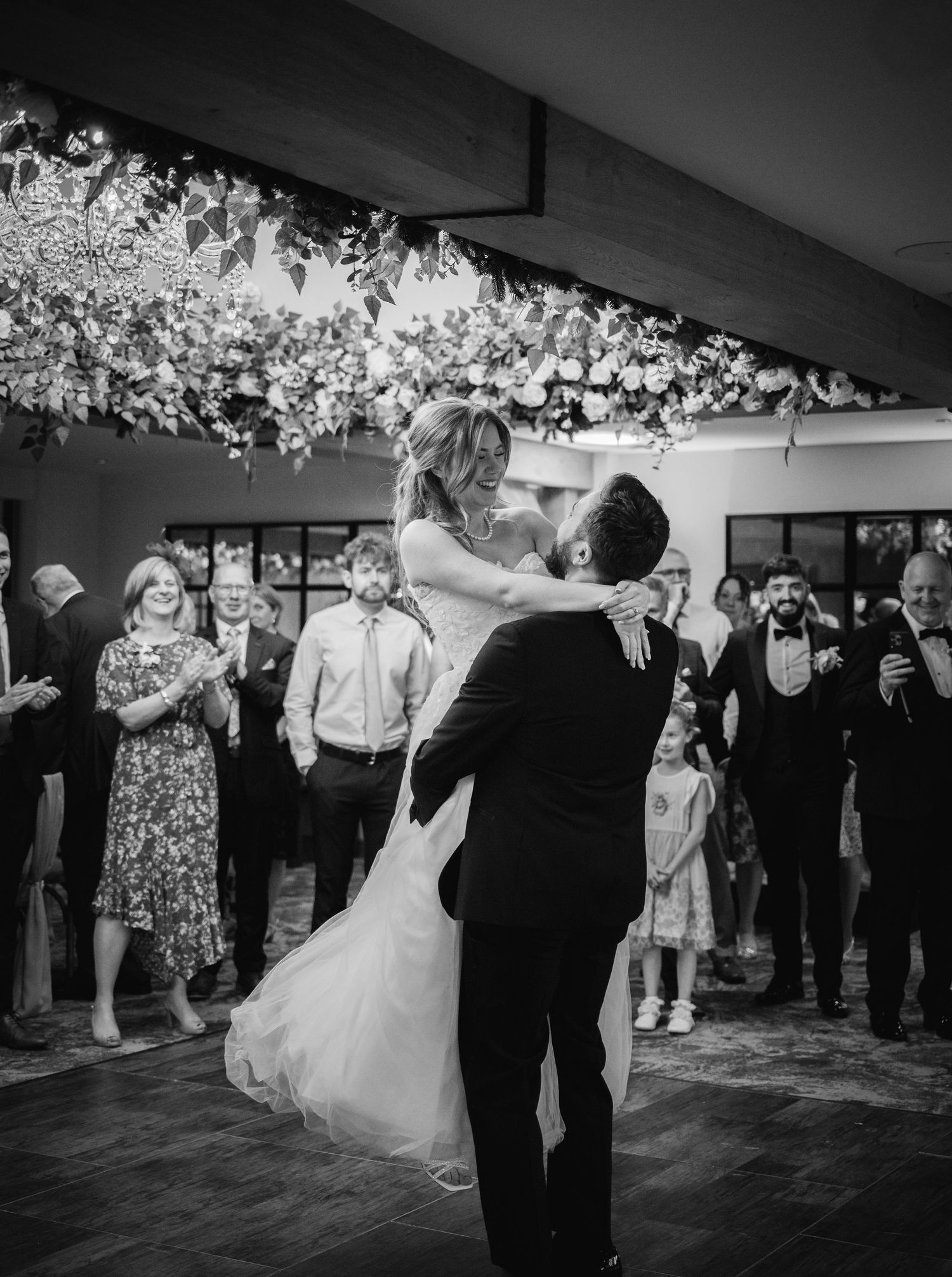 Bride and groom share first dance surrounded by guests under floral ceiling installation at elegant wedding reception.