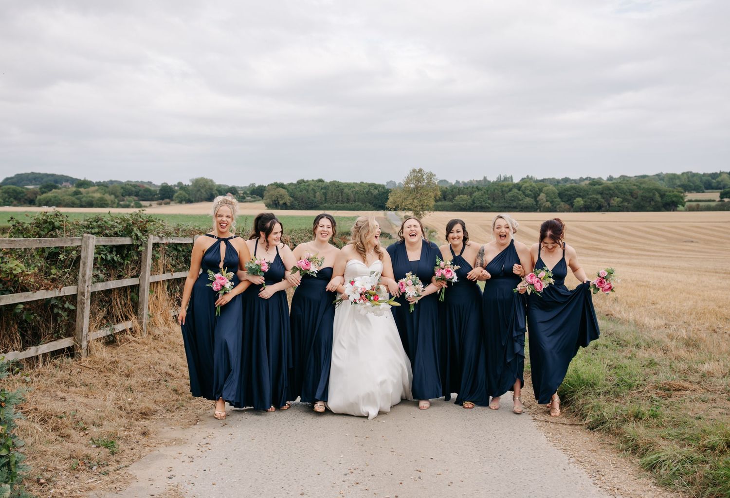 Wedding party in navy blue dresses and white gown poses together along a wooden fence in an open field.