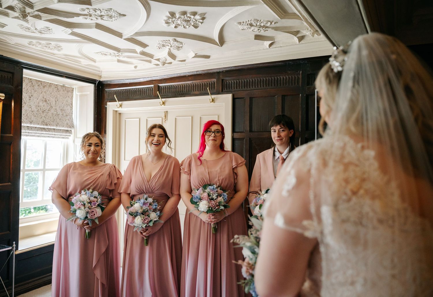 Bridesmaids in pink dresses smile as bride in white lace gown and veil reveals her wedding look indoors.