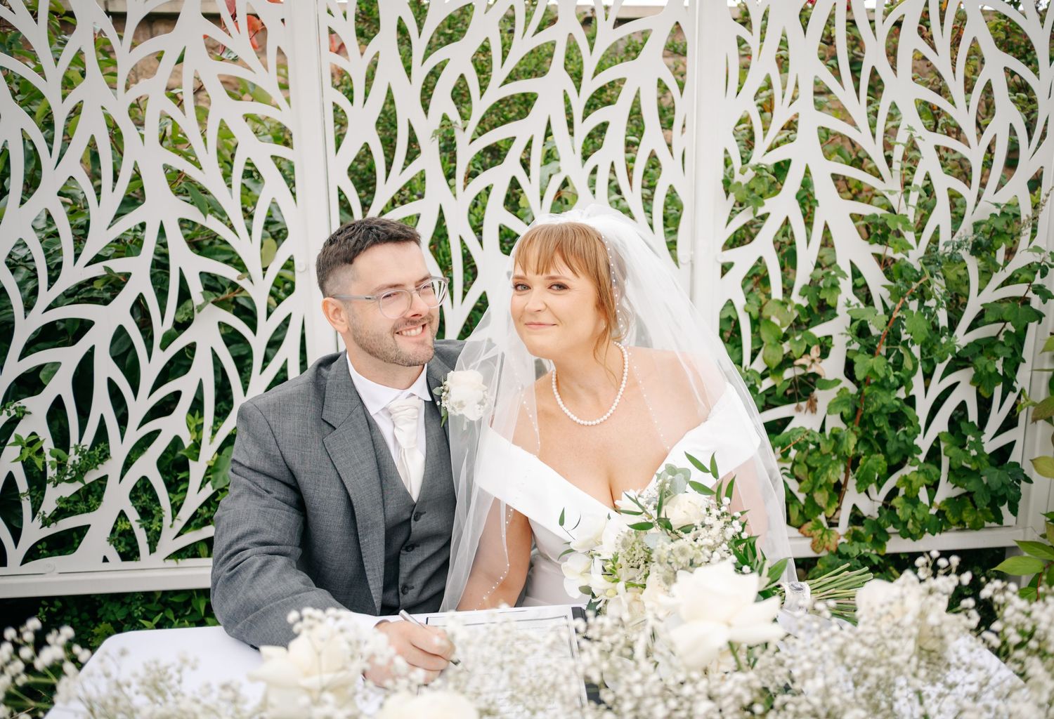 Wedding couple seated at decorated table with white floral arrangements and greenery.