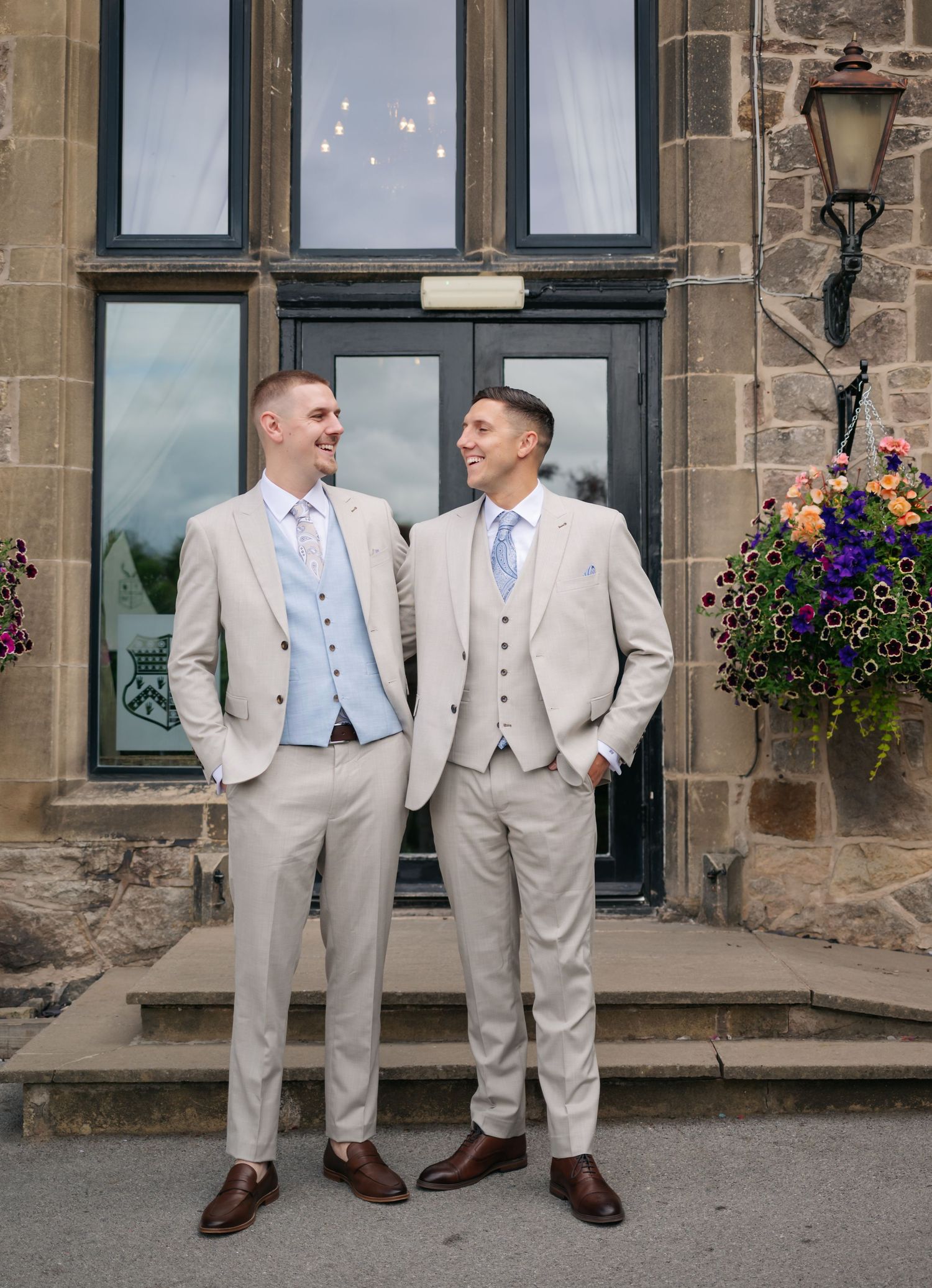 Groomsmen in light beige suits and blue ties pose together against a stone building with hanging flower baskets.