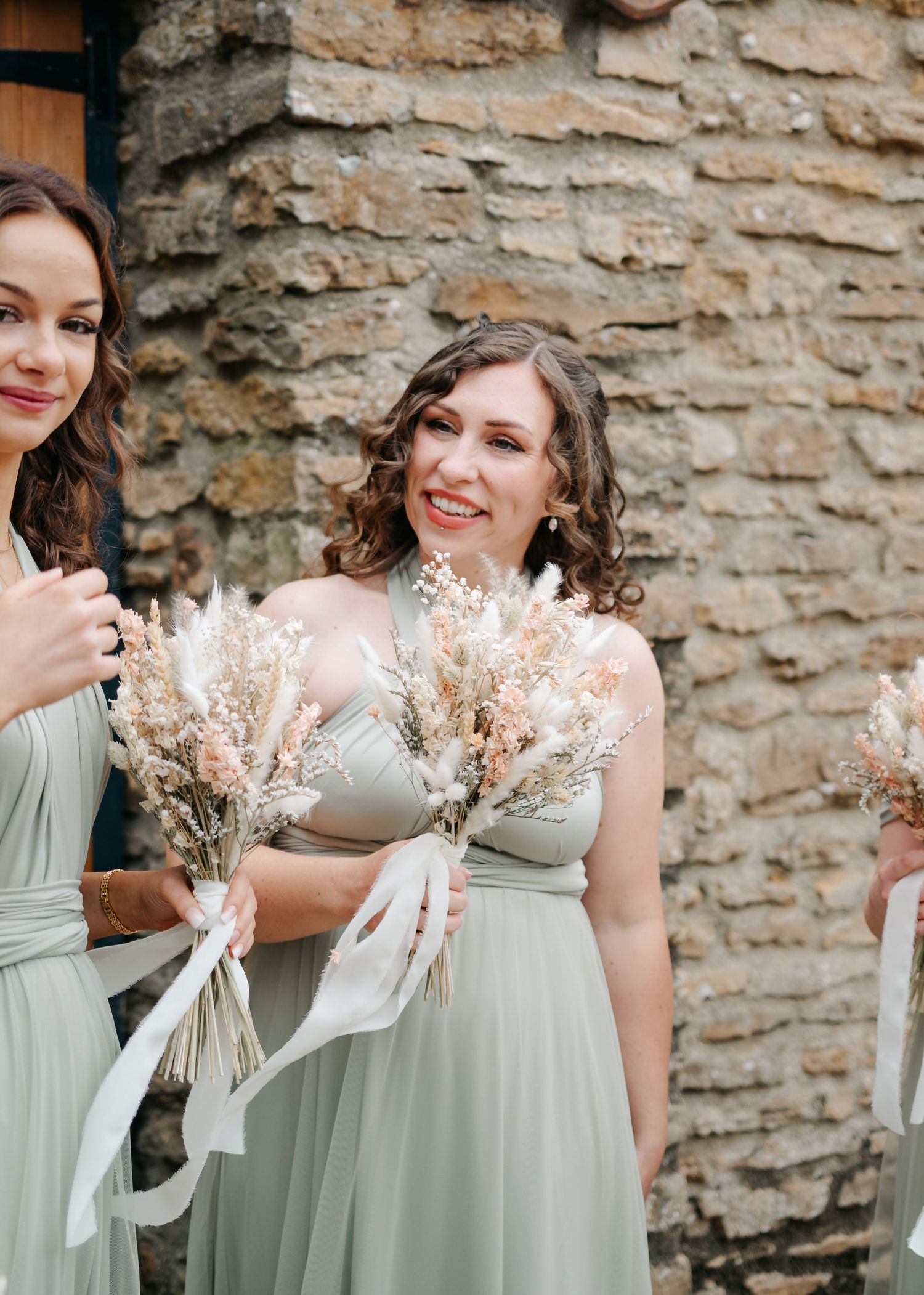 Bridesmaids in sage green dresses holding dried flower bouquets against stone wall.