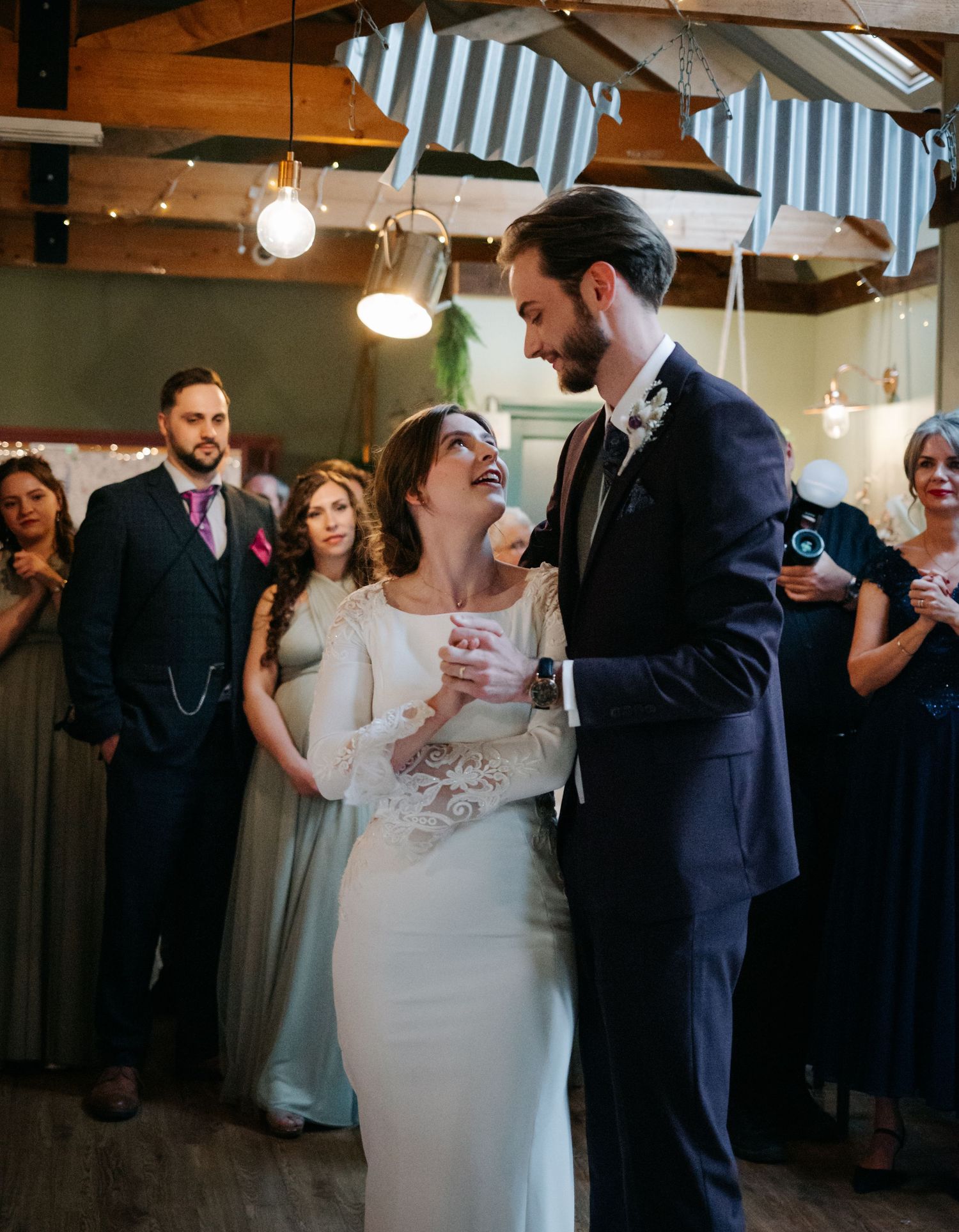 Wedding party dancing together under string lights in rustic indoor venue.