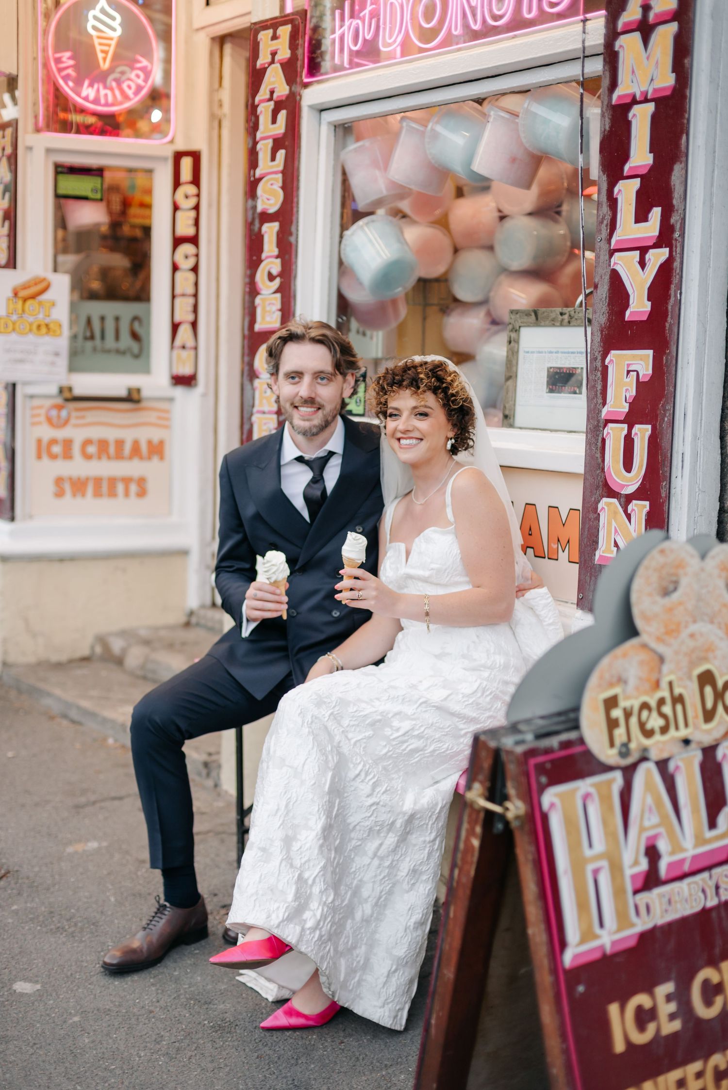 Newlyweds share ice cream outside vintage shop with pink neon signs and pastel decorations.
