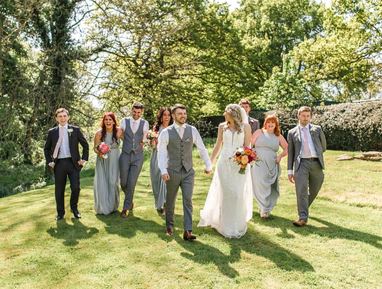 Wedding party walking and laughing together on grassy lawn during outdoor celebration.