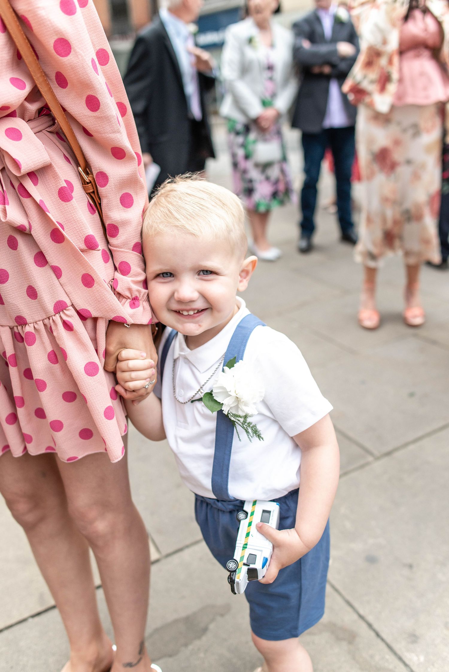 A young groomsman in navy shorts and white shirt holds a toy while standing next to a polka dot dress.