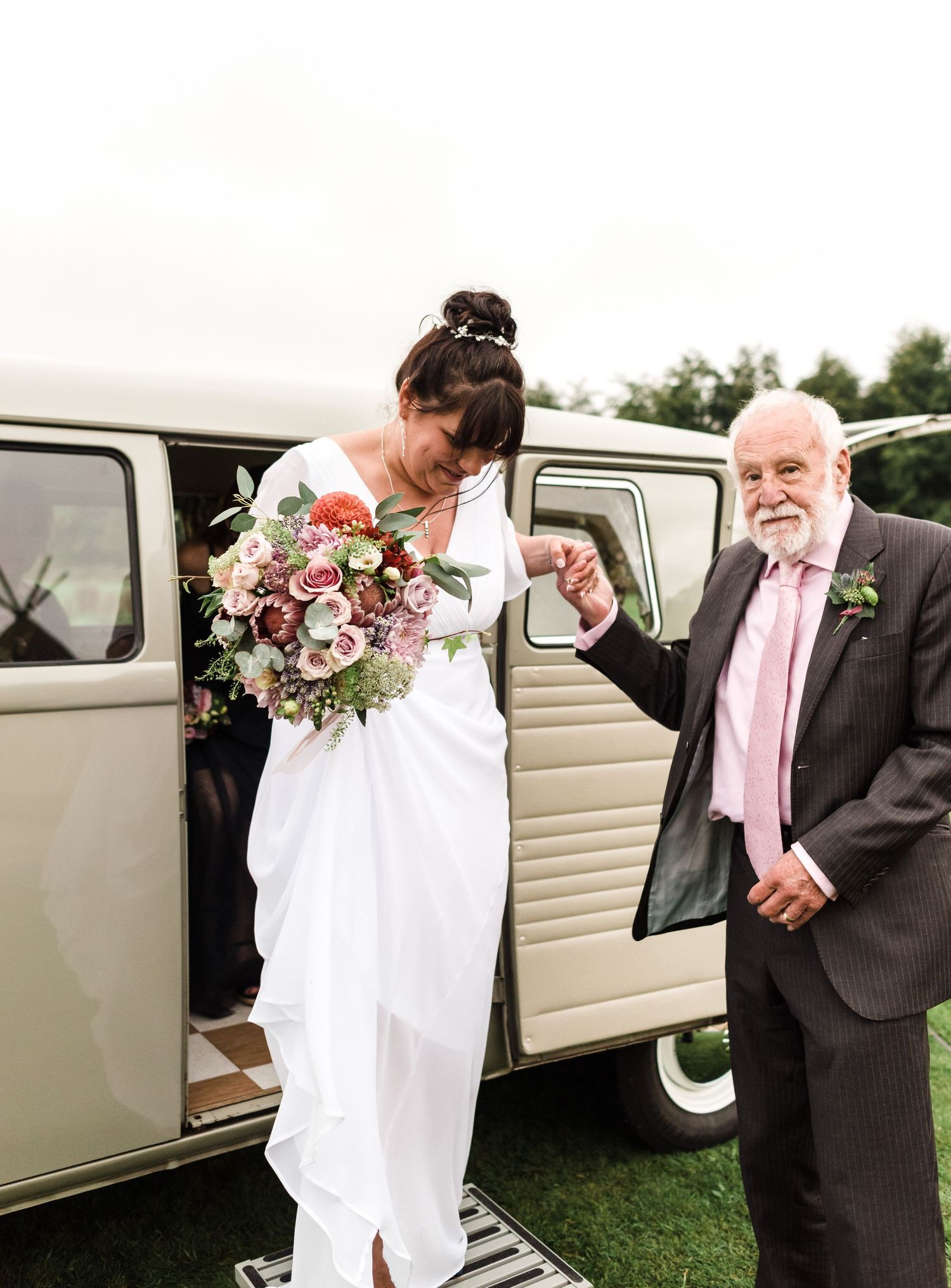 A vintage cream-colored VW bus serves as wedding transportation on a grassy field.