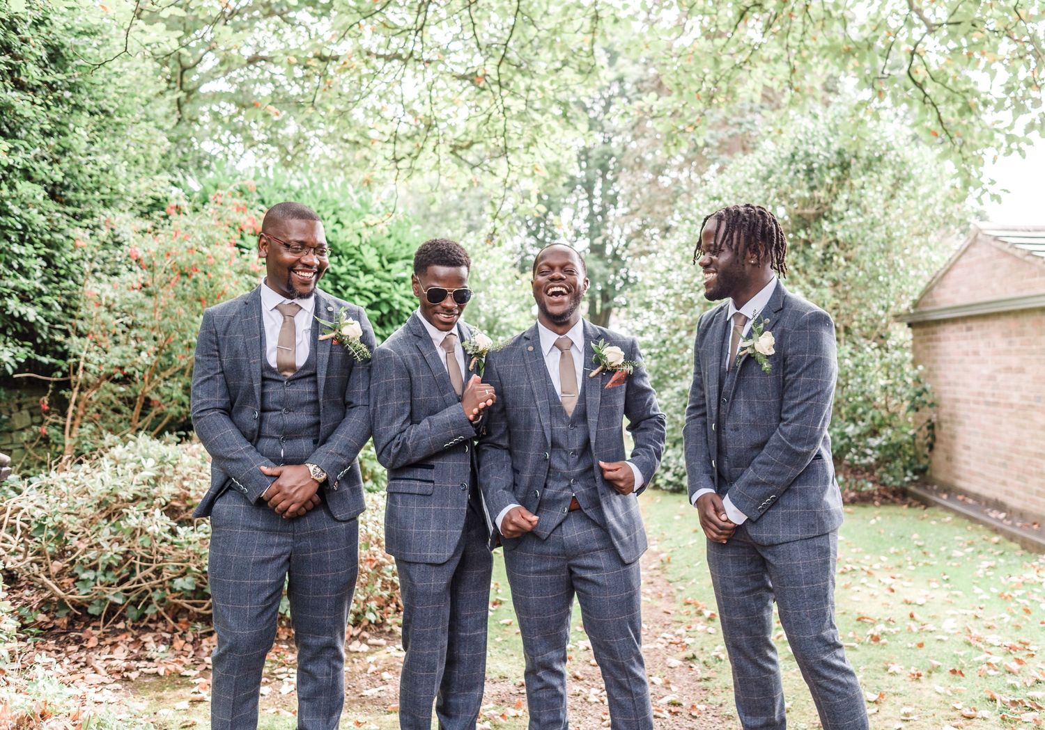 Four groomsmen in coordinating gray suits pose together in a garden setting.